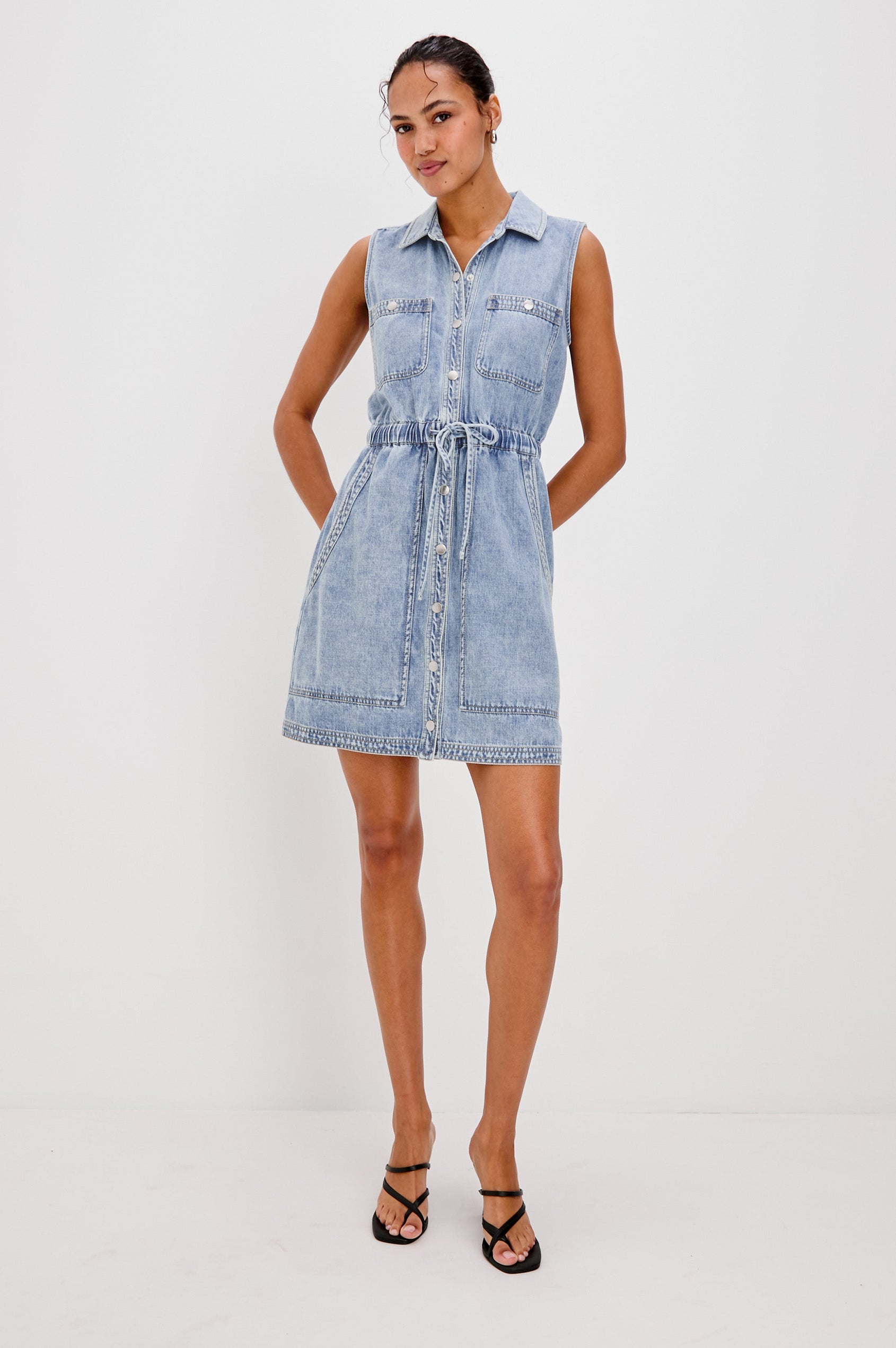 Erica Lightweight Denim Mini Dress with a Exposed Button Front, Encased Drawstring Waistband, Front Patch Pockets, Chest Pockets, Sleeveless and Collared in Faded Indigo Colorway. Full Body Front View Shown on Model.