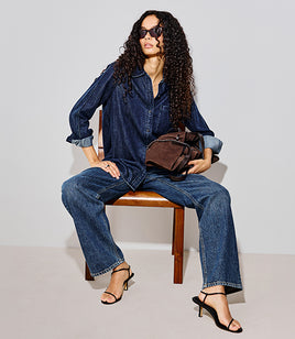 A model with long curly dark hair sits on a wooden chair wearing a matching blue denim long-sleeve button-up shirt and wide-leg jeans, holding a brown leather bag, and wearing black strappy high-heeled sandals against a light gray studio background.