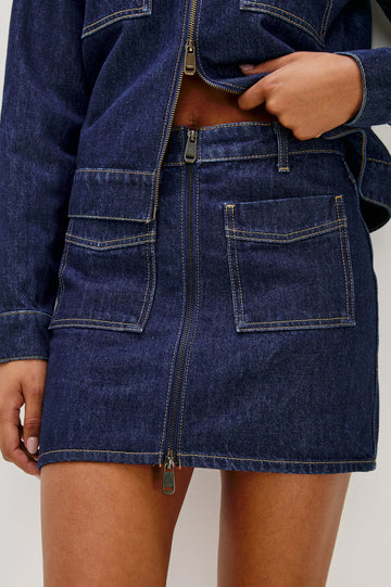 The Fallbrook is a mini zip front denim skirt with beltloops, front patch pockets and back pockets in Deep Indigo colorway. Front detail view featured on model.