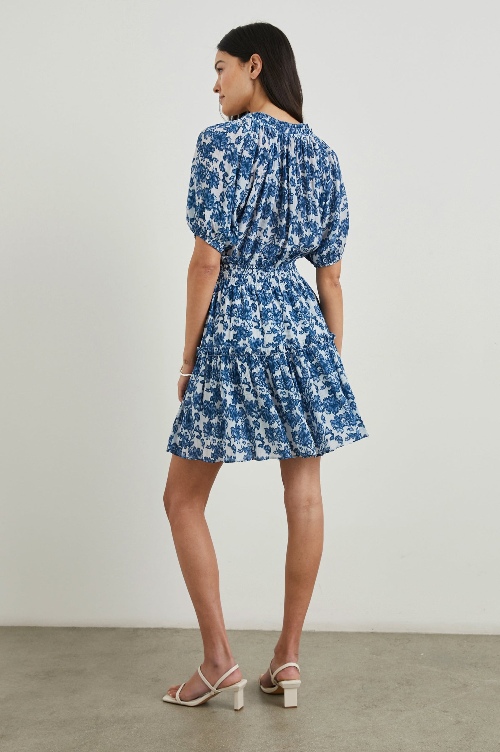 Fiorella Short-Sleeve, Above-Knee Dress with a Shirred Collar and Waist, V-neckline with Ties, Puff Sleeves and a Tiered Skirt in Chambray Floral Colorway,  Blue Flowers on White - Back Full Body View Featured on Model
