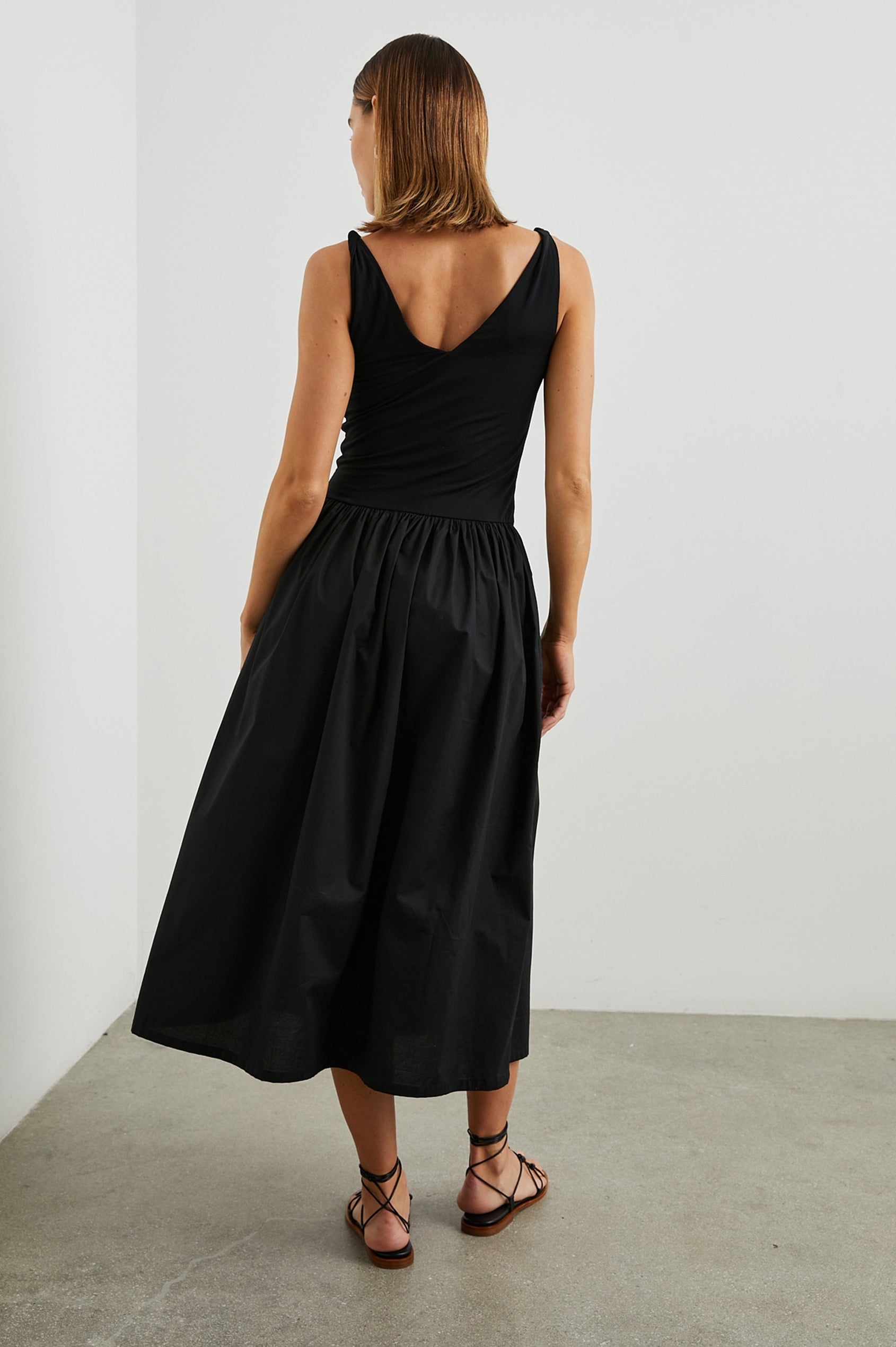 Franca Sleeveless Midi Dress with Twisted Shoulder Straps, a deep V Neck and Pleats at the Waist in Black Colorway - Back Full Body View Featured on Model