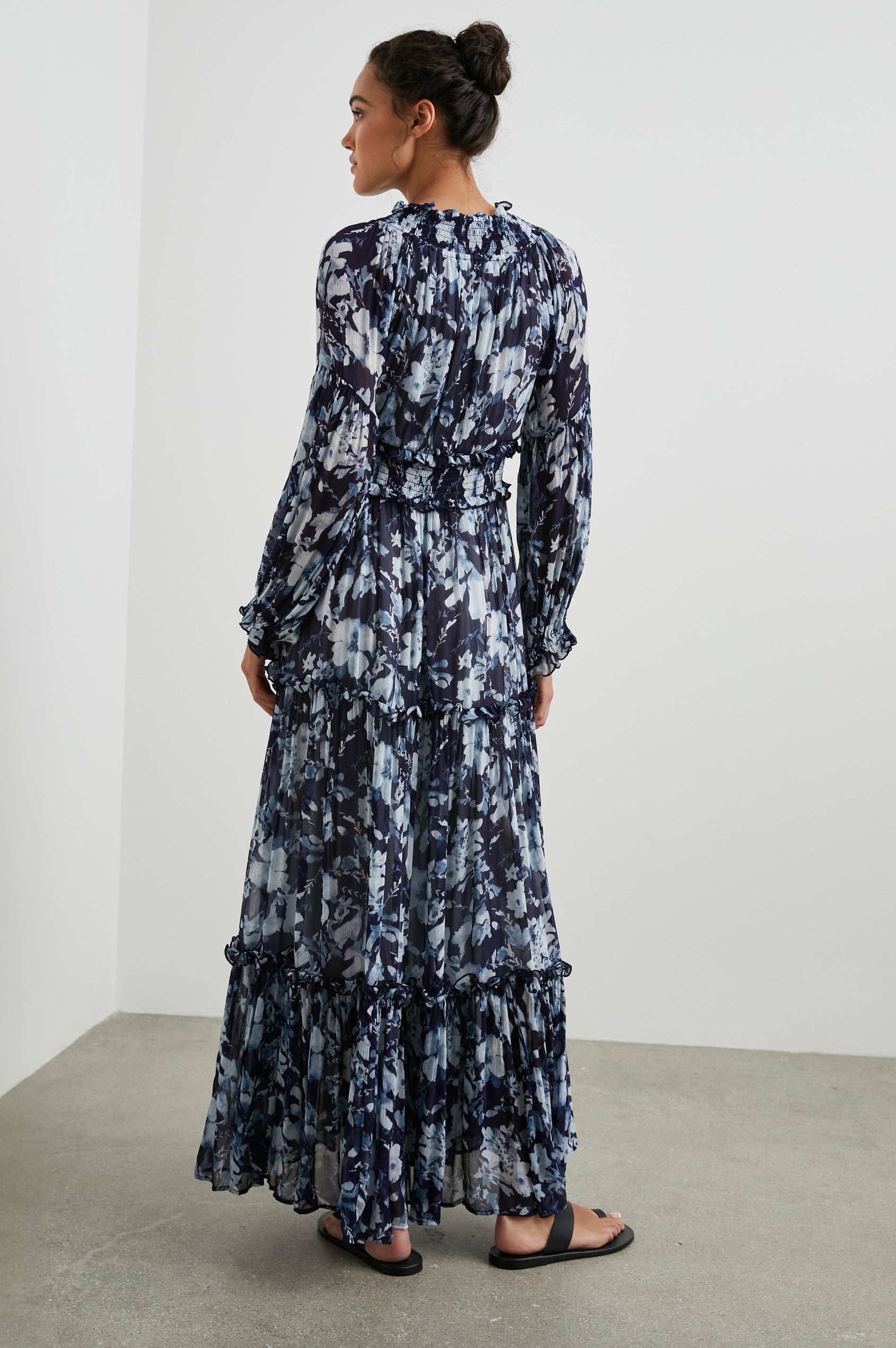 Frederica Long-Sleeve Maxi Dress with a V Neck, a Shirred Collar and Waist, Elastic Cuffs and a Tiered Skirt in Indigo Blossoms Colorway, with blossoms in Blues on Indigo - Back Full Body View Featured on Model