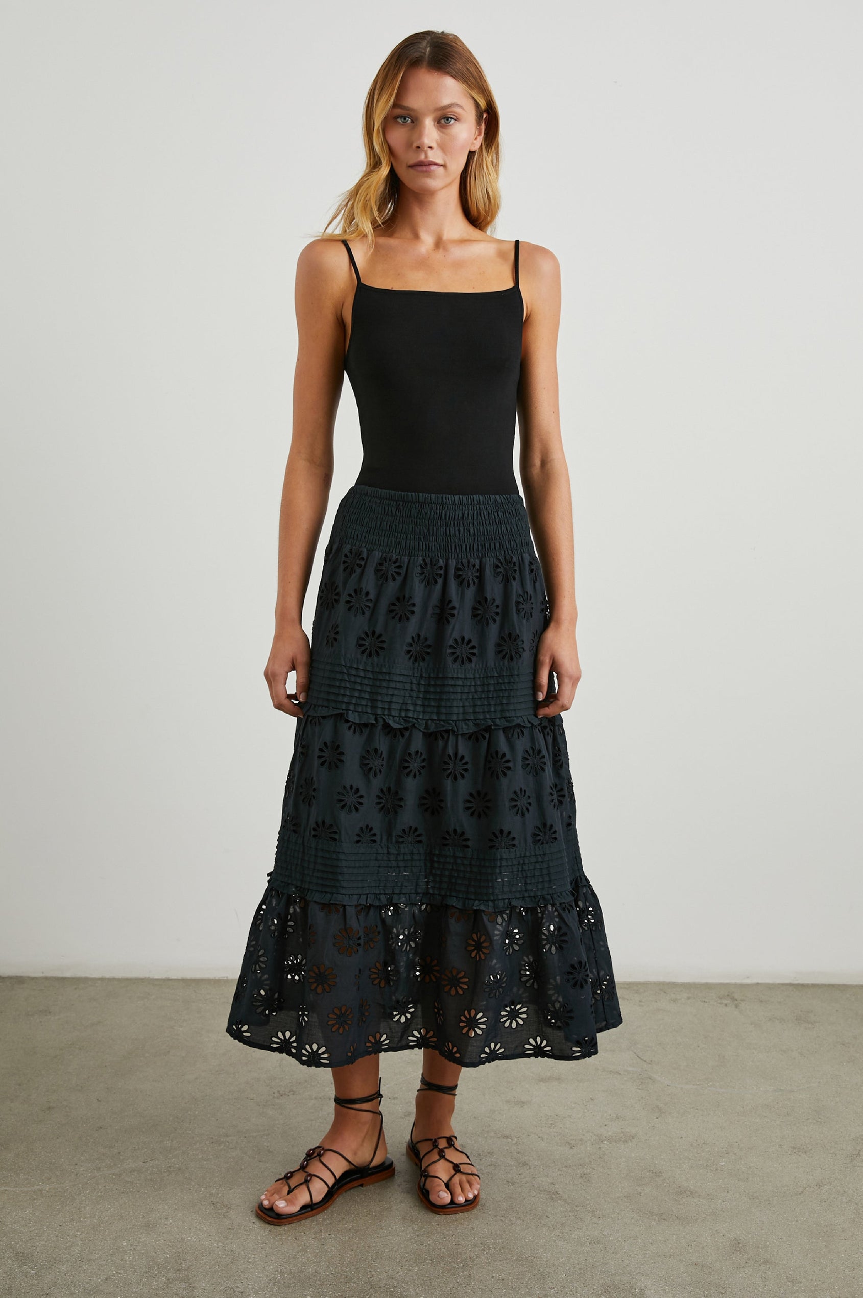 Gail Tiered Midi Skirt with a Shirred Waist, Horizontal Pleating and Ruffles, and Eyelet Flowers in Black Colorway - Front Full Body View Featured on Model 
