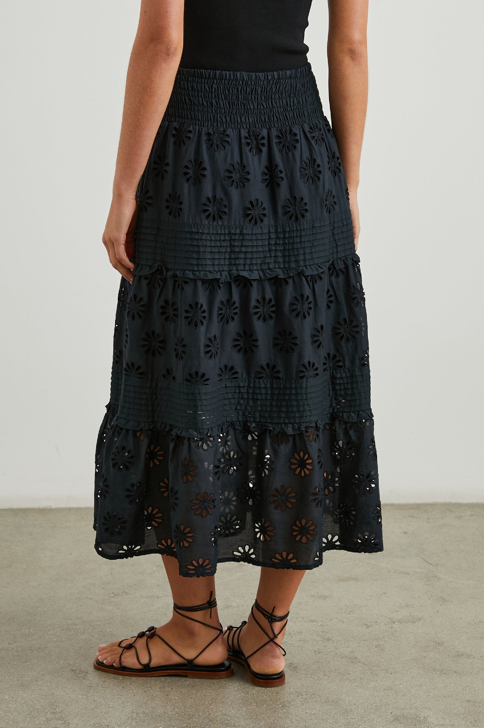 Gail Tiered Midi Skirt with a Shirred Waist, Horizontal Pleating and Ruffles, and Eyelet Flowers in Black Colorway - Back View Featured on Model 