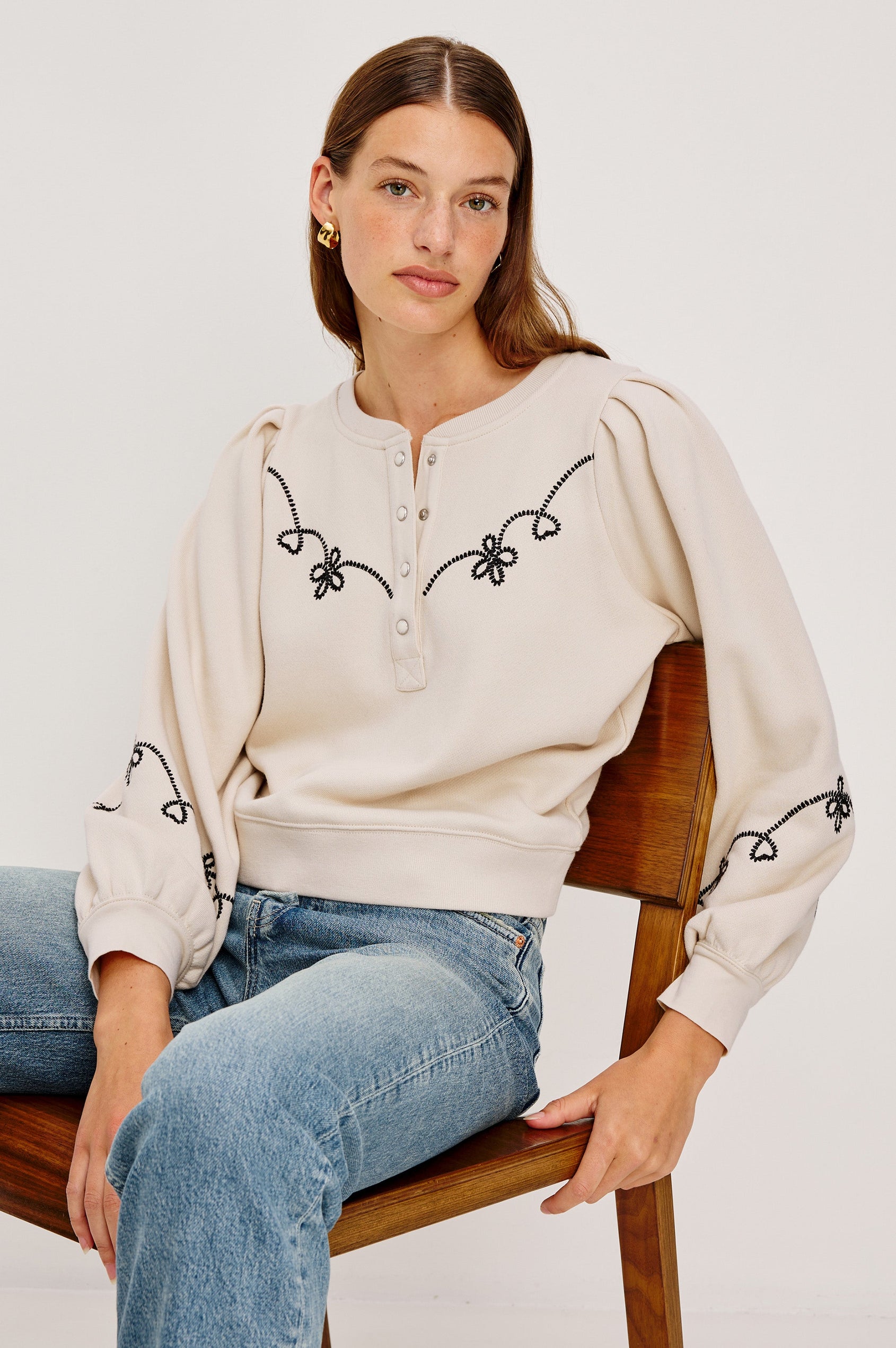 The Hanne is a henley style long sleeve sweatshirt with a snap front placket, rib trim, western style contrast black embroidery and puff sleeves in Ecru colorway. Front seated view featured on model.