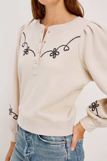 The Hanne is a henley style long sleeve sweatshirt with a snap front placket, rib trim, western style contrast black embroidery and puff sleeves in Ecru colorway. Front detail view featured on model.