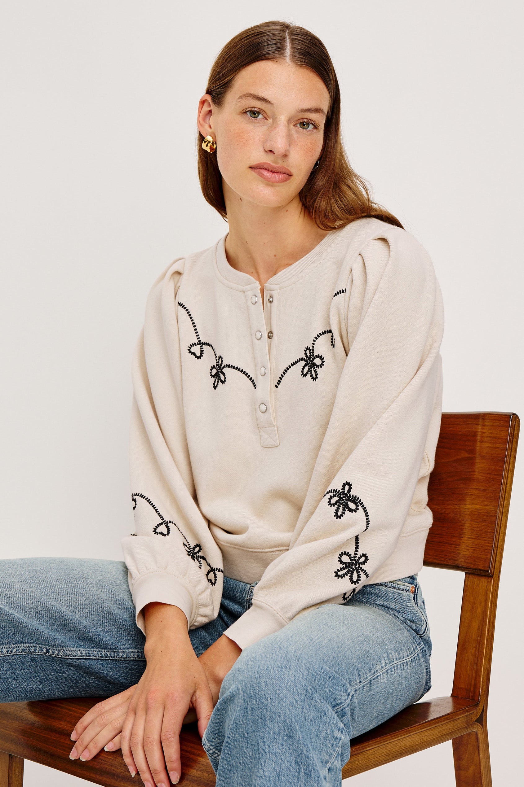 The Hanne is a henley style long sleeve sweatshirt with a snap front placket, rib trim, western style contrast black embroidery and puff sleeves in Ecru colorway. Front seated view featured on model.