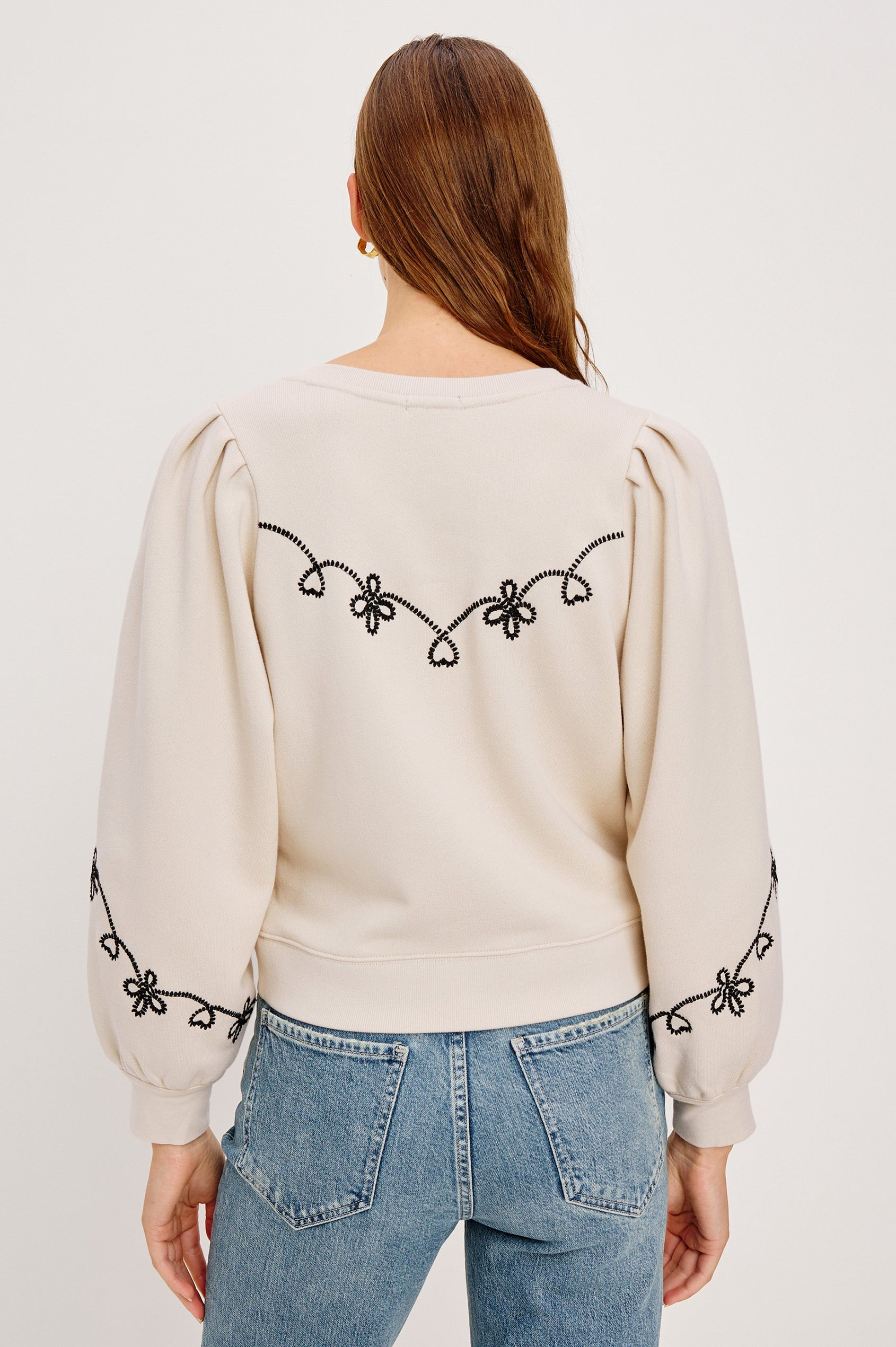 The Hanne is a henley style long sleeve sweatshirt with a snap front placket, rib trim, western style contrast black embroidery and puff sleeves in Ecru colorway. Back seated view featured on model.
