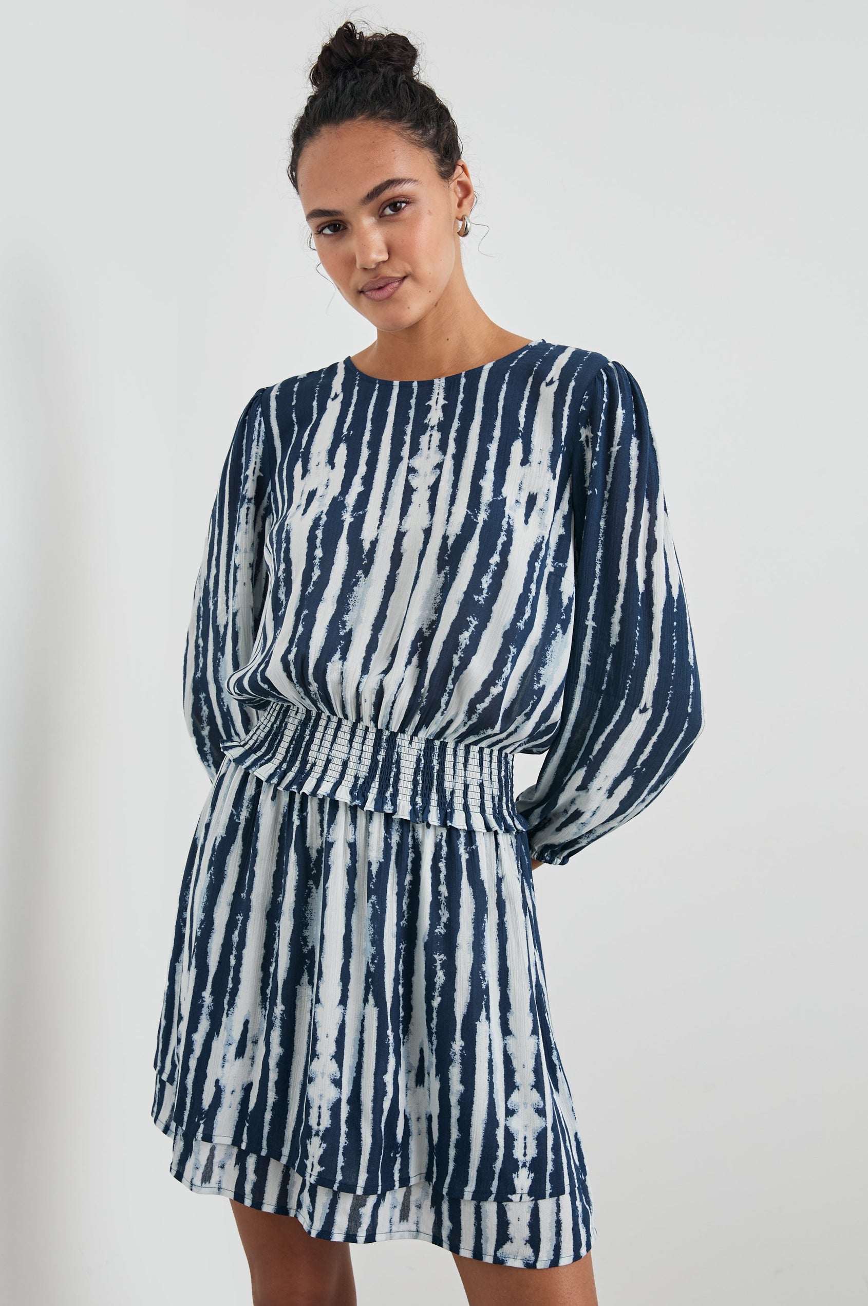 Inez Mini Dress with Three-Quarter Sleeves, a Shallow Scoop Neck, a Smocked Waist, Elasticized Cuffs and a Double Layer Skirt in Indigo Nile Colorway, White and Blue Vertical Streaks - Front View Featured on Model 
