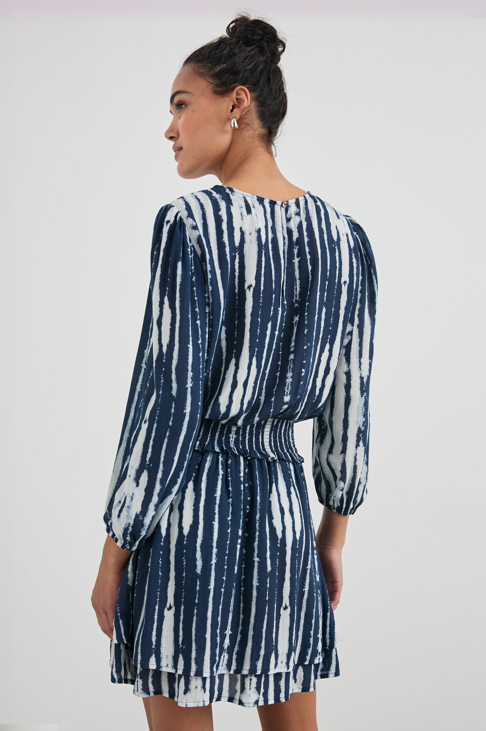 Inez Mini Dress with Three-Quarter Sleeves, a Shallow Scoop Neck, a Smocked Waist, Elasticized Cuffs and a Double Layer Skirt in Indigo Nile Colorway, White and Blue Vertical Streaks - Back View Featured on Model 