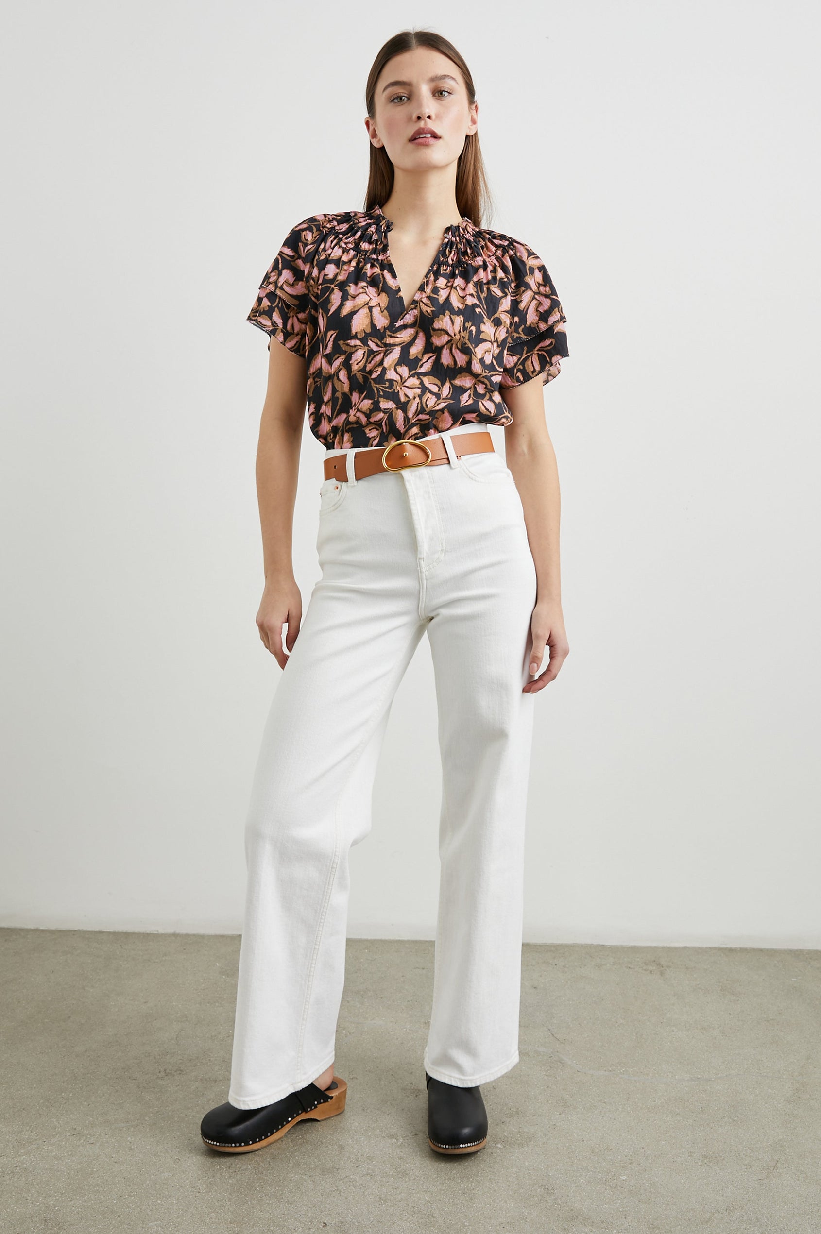 Kamila Short-Sleeve Top with Ruffled Sleeves, Deep V neck, and a Shirred Collar with a button in Jasmine Colorway, with pink and brown flowers on black - Front Tucked Full Body View Featured on Model