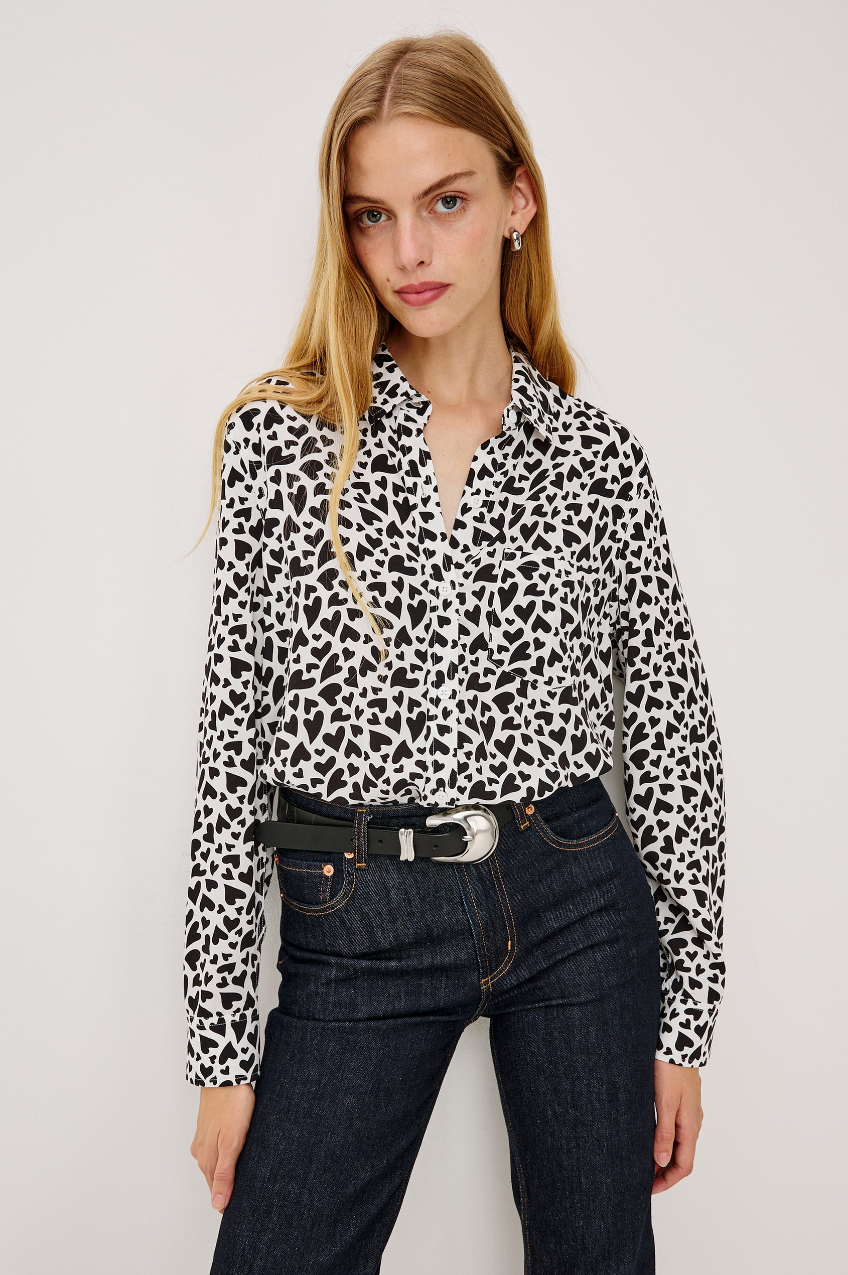 A white, long-sleeved button-down shirt featuring an all-over print of small black hearts, styled with a classic collar and a single chest pocket - front crop view on model
