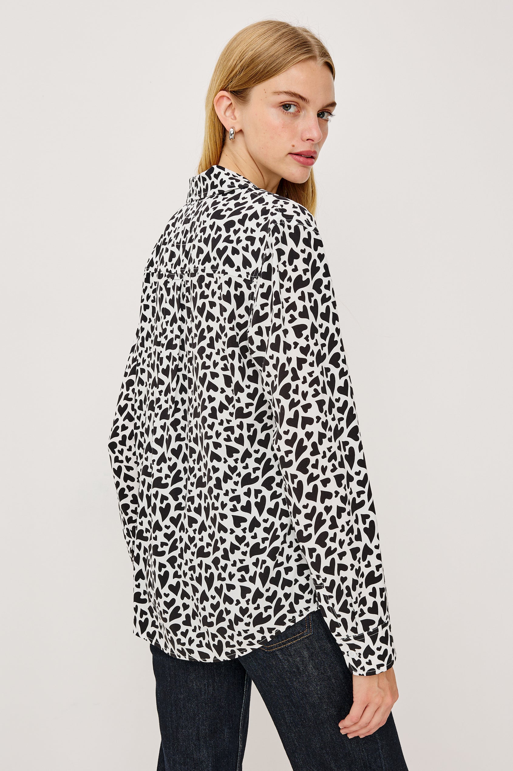 A white, long-sleeved button-down shirt featuring an all-over print of small black hearts, styled with a classic collar and a single chest pocket - back crop view on model
