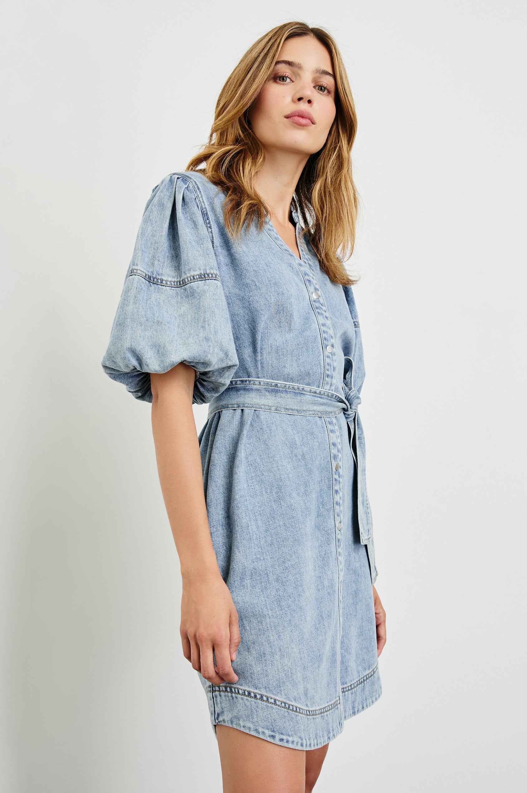 Kingsley Denim Mini Dress with Elbow-Length Puff Sleeves, a V Neck, Snaps Down the Front and a Fabric Tie Belt in Faded Indigo Colorway - Side View Featured on Model 