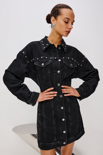 Ladera is a trucker style denim mini dress with long sleeves, drop shoulders, relaxed balloon sleeves, nail-back studs, and front button closures, with a subtle A-line shape and adjustable back strap in Black Stone Studded colorway. Front view featured on model.
