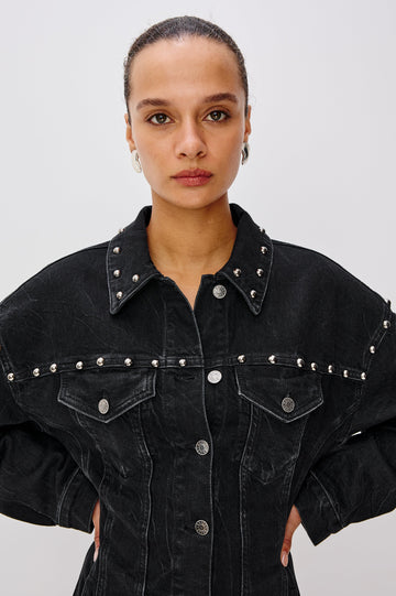 Ladera is a trucker style denim mini dress with long sleeves, drop shoulders, relaxed balloon sleeves, nail-back studs, and front button closures, with a subtle A-line shape and adjustable back strap in Black Stone Studded colorway. Front detail view featured on model.
