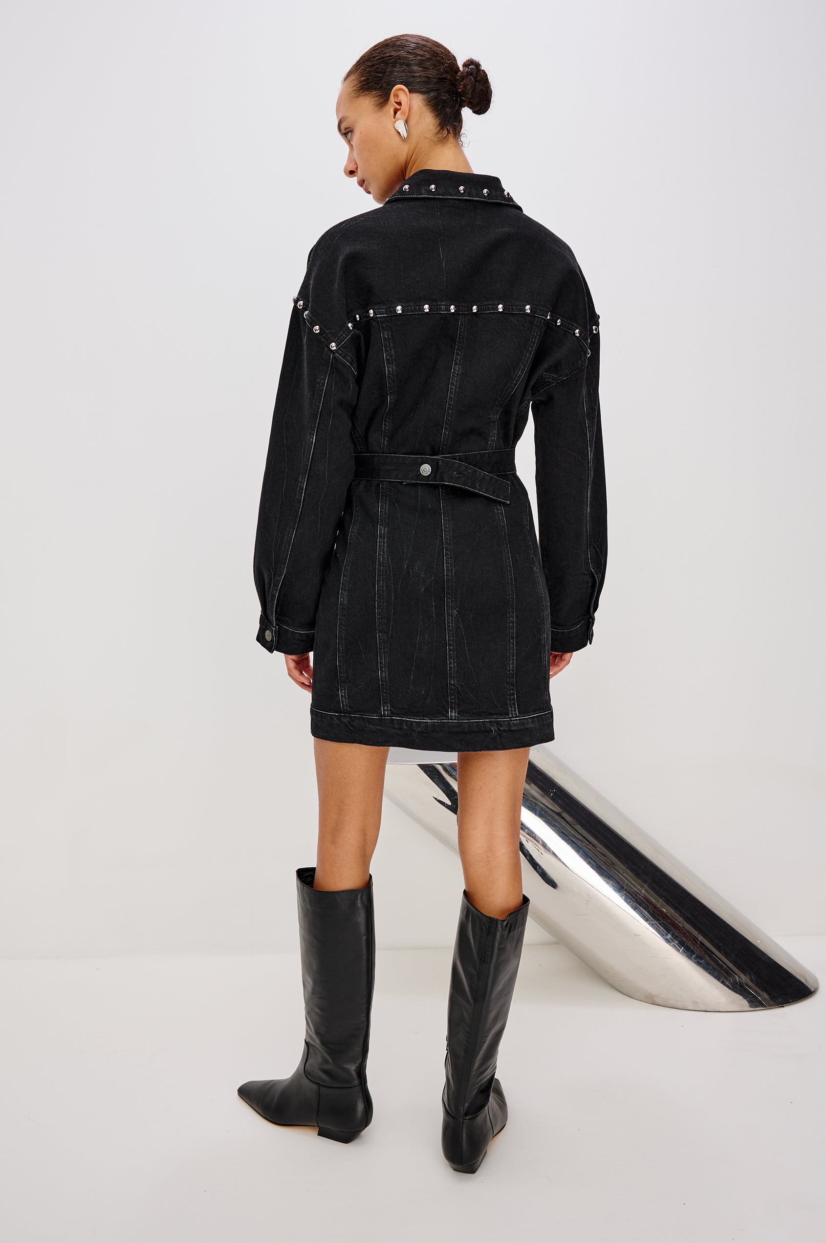 Ladera is a trucker style denim mini dress with long sleeves, drop shoulders, relaxed balloon sleeves, nail-back studs, and front button closures, with a subtle A-line shape and adjustable back strap in Black Stone Studded colorway. Back full body view featured on model.