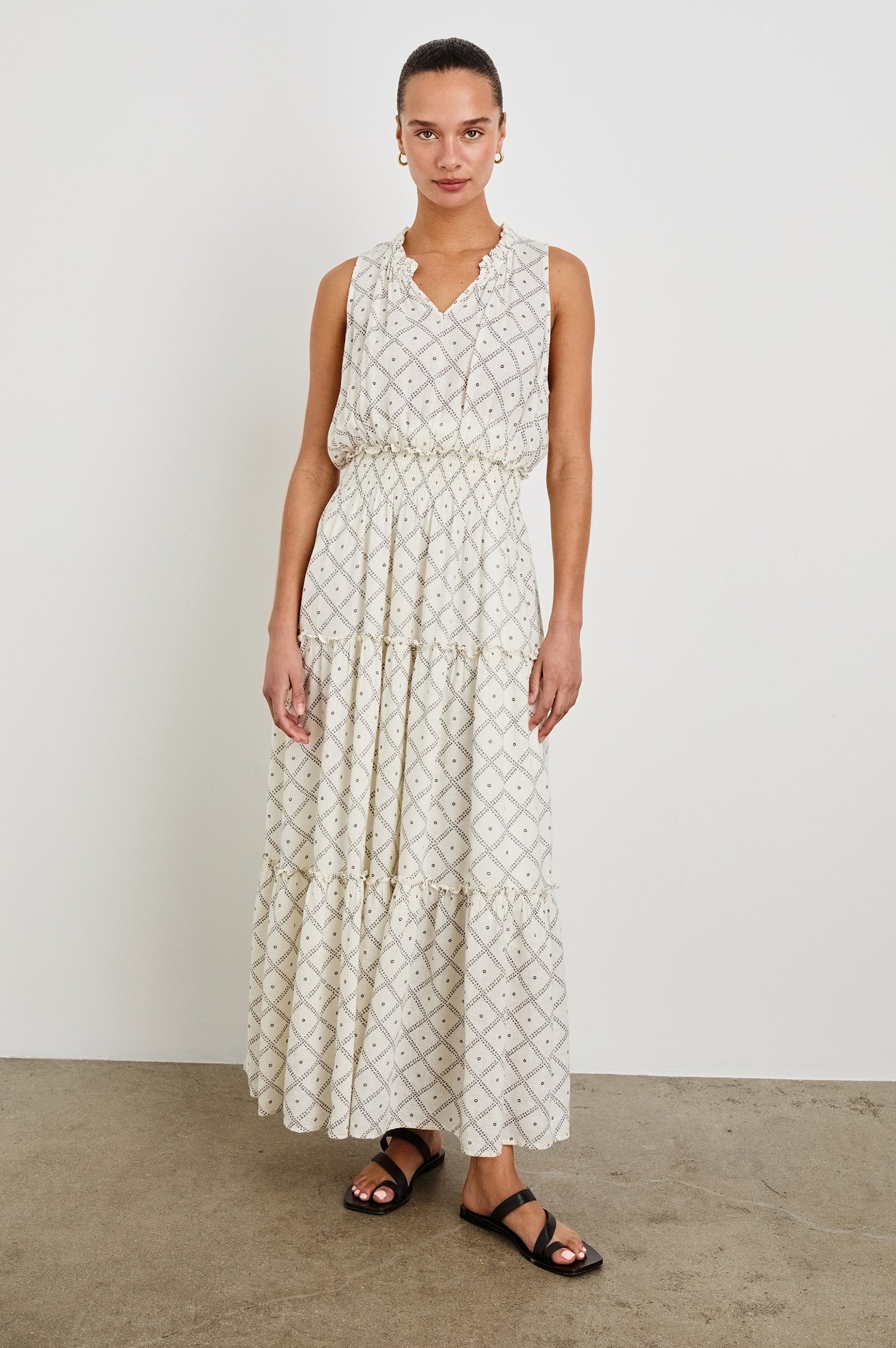 Female model is wearing the Loulou Maxi Dress in a black and white boho print. Dress is Ivory with black printed details. Features smocking at the waist, a v-neckline and fully-lined skirt with ruffle detail at each tier.