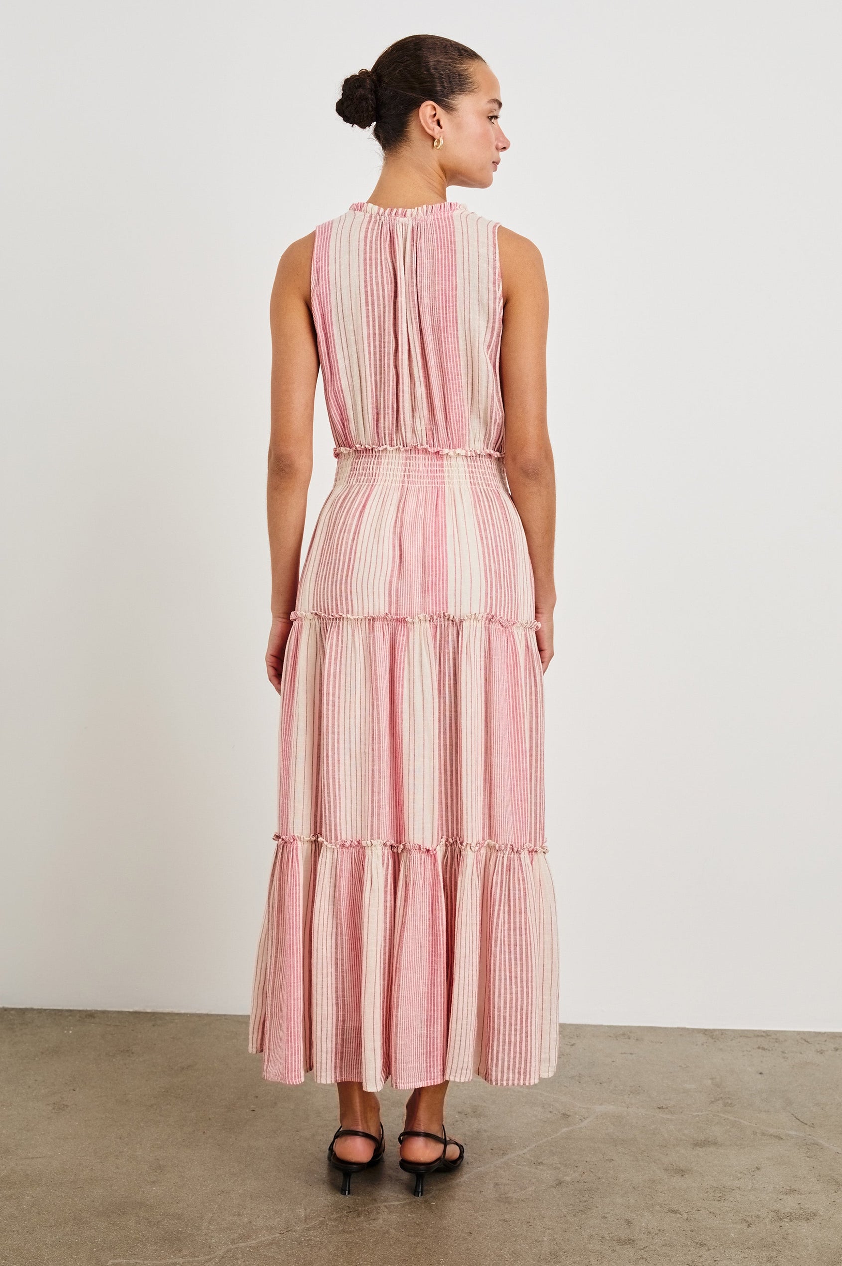Back view of female model wearing our sleeveless, Loulou Maxi Dress in color Salerno Stripe, a pink and white boho stripe. Dress features smocked waist, v-neck, and tiered maxi skirt. 
