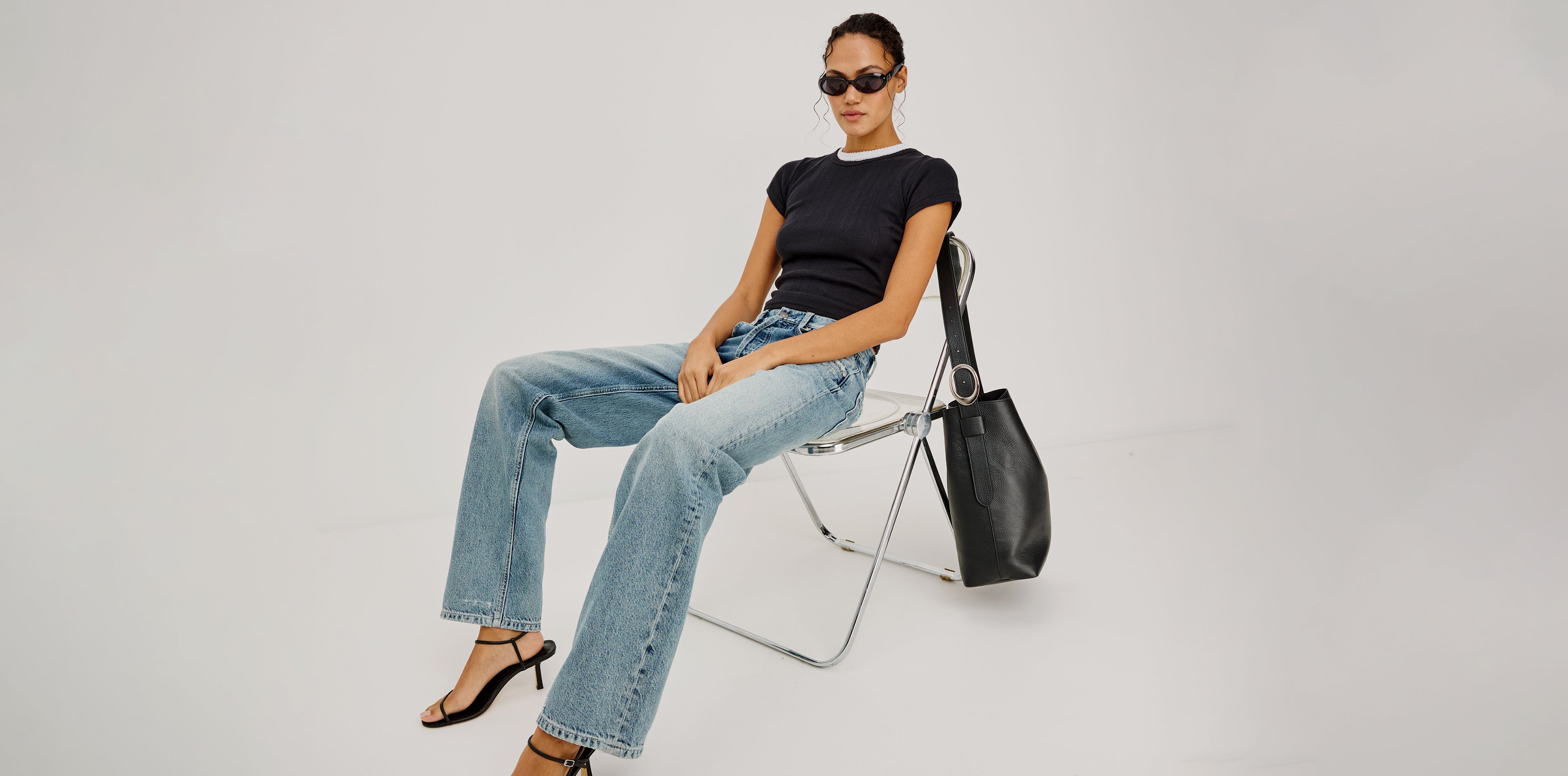 Woman sitting on a chair wearing a black t-shirt, blue jeans, and black high-heeled sandals with a black handbag.