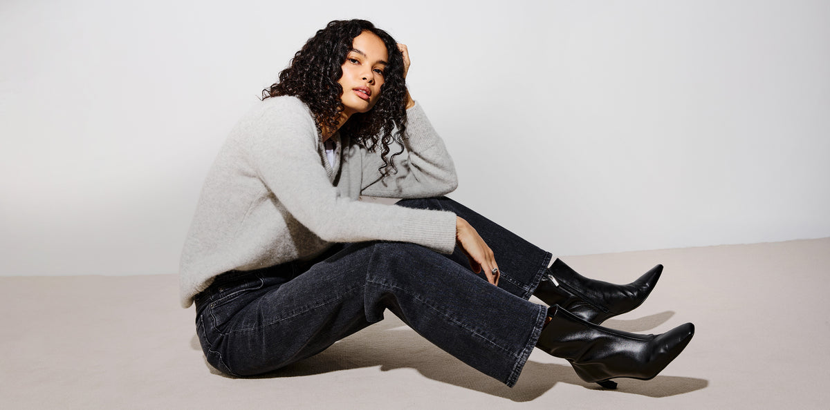 A woman sits cross-legged on a light beige floor against a white studio wall, wearing a light gray knit sweater, dark wide-leg jeans, and black ankle boots.