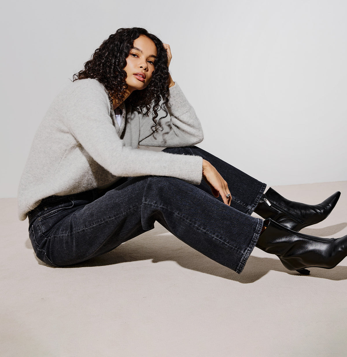 A model sits on a light beige floor against a white studio background, wearing a light gray knit sweater, dark wide-leg jeans, and black ankle boots.