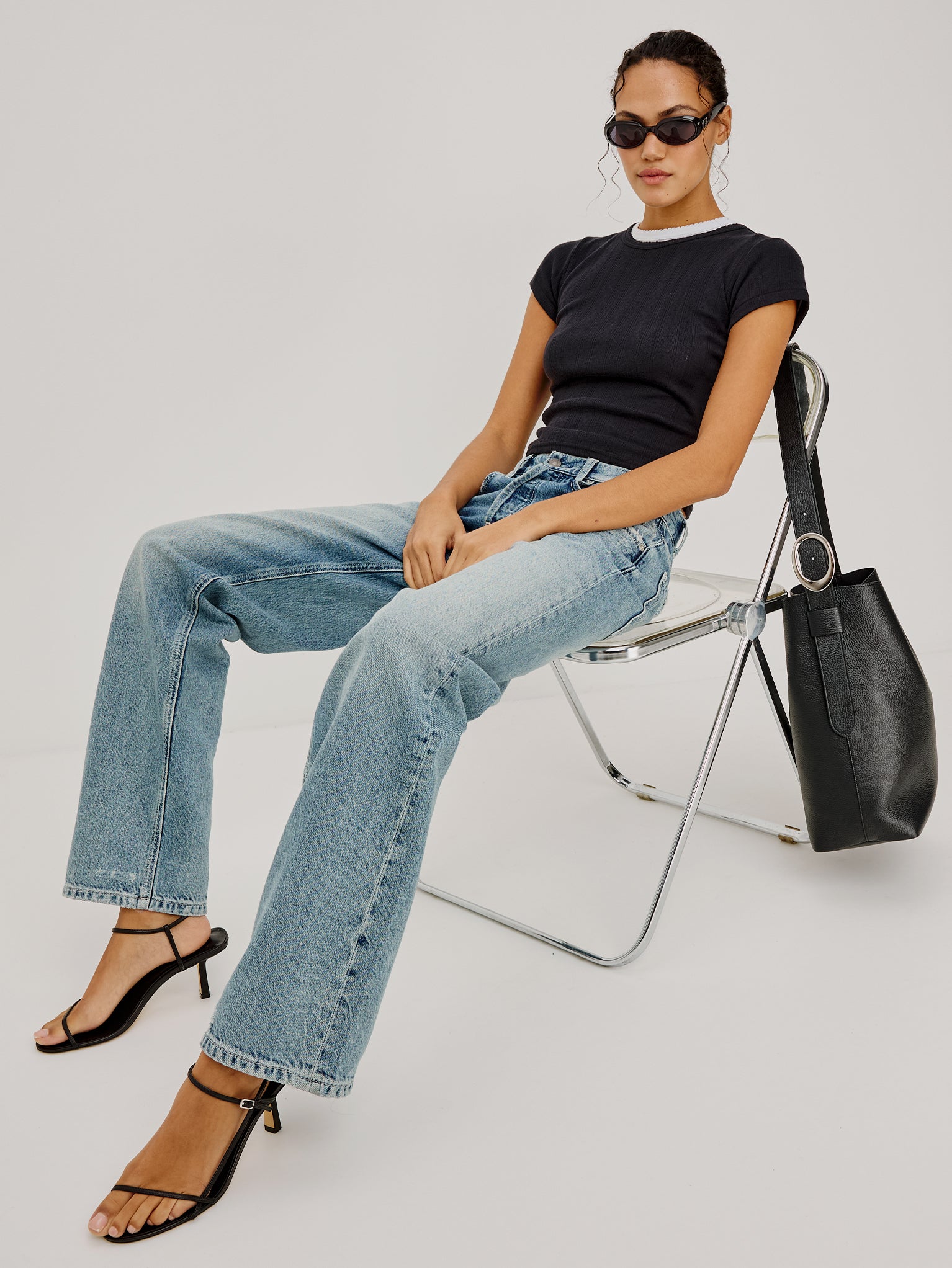 Woman sitting on a chair wearing a black t-shirt, blue jeans, and black high-heeled sandals with a black handbag.