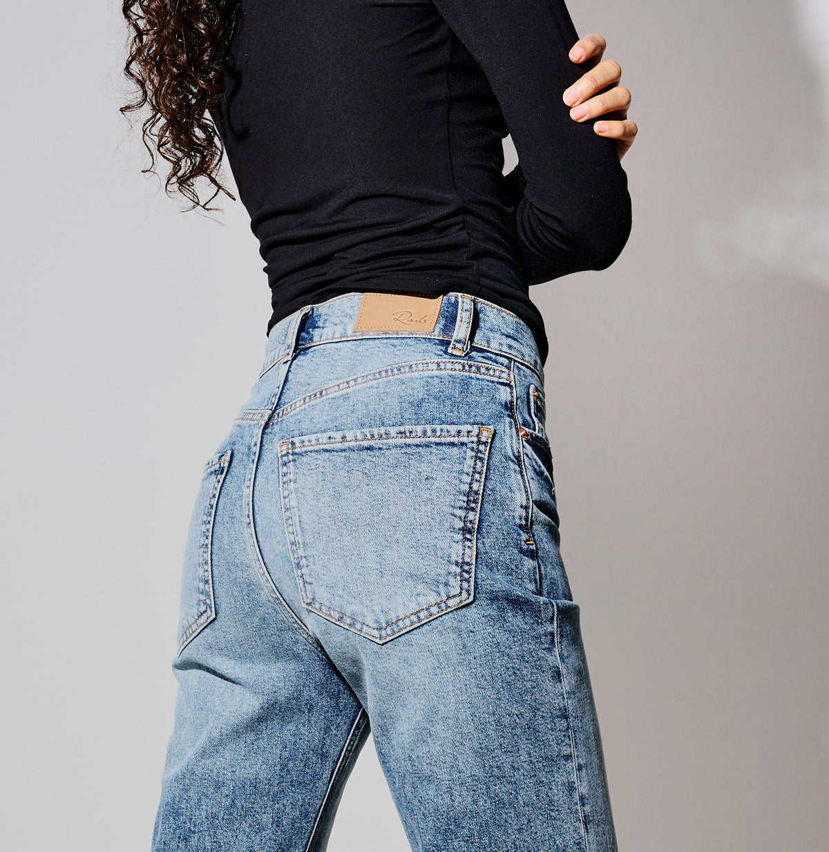 Rear view of a person wearing light-wash blue denim jeans and a fitted black long-sleeve top against a plain light gray studio background.