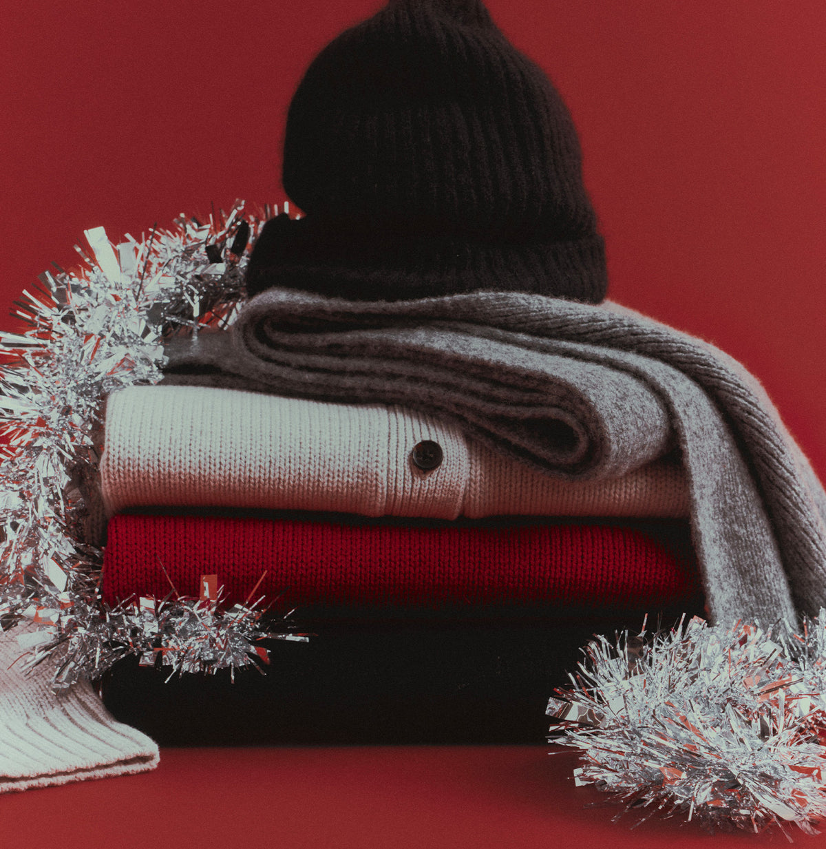 A stack of folded knit winter garments and a black ribbed beanie arranged with silver tinsel on a red background. Visible items include a light gray carded sweater, a red knit layer, a dark garment, and a cream knit sleeve or mitt.