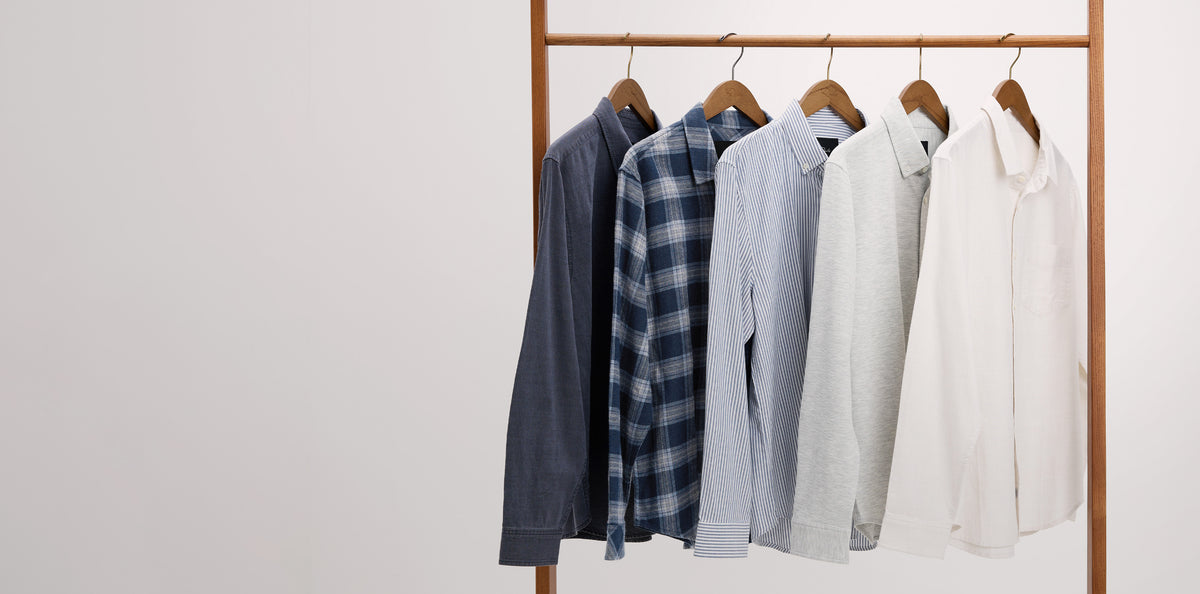 Five long-sleeve button-up shirts in different colors and patterns hang on a wooden clothing rack against a plain white wall. The shirts include dark navy, blue plaid, light blue, light gray, and off-white.