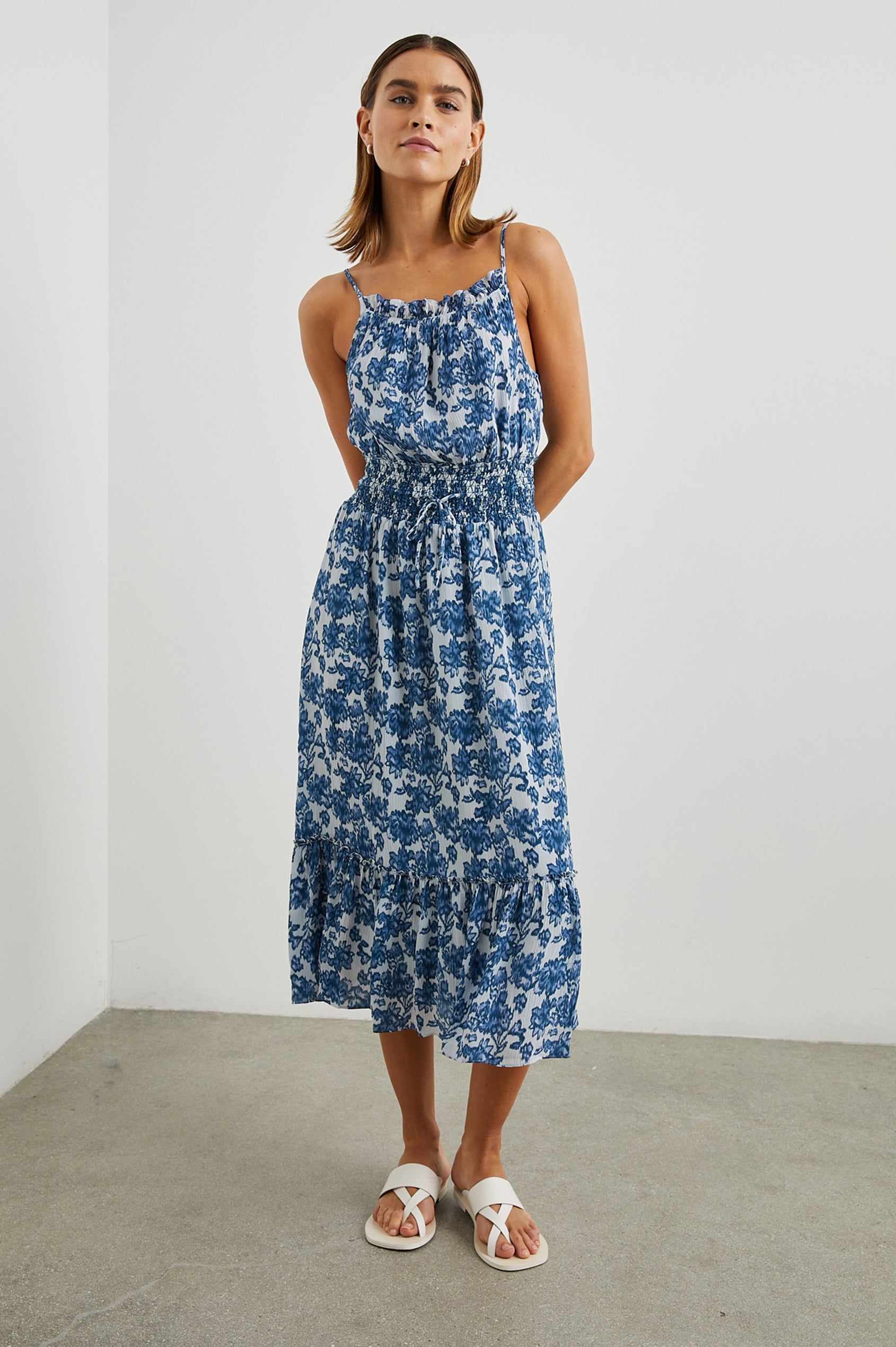 Magdalene Sleeveless Midi Dress with Spaghetti Straps, a Button Keyhole at the Back, a Shirred Waist with a Drawstring, a Scoop Neckline with Ruffled Trim, and a Ruffled Hem in Chambray Floral Colorway, Blue Flowers on White - Front Full Body View Featured on Model  