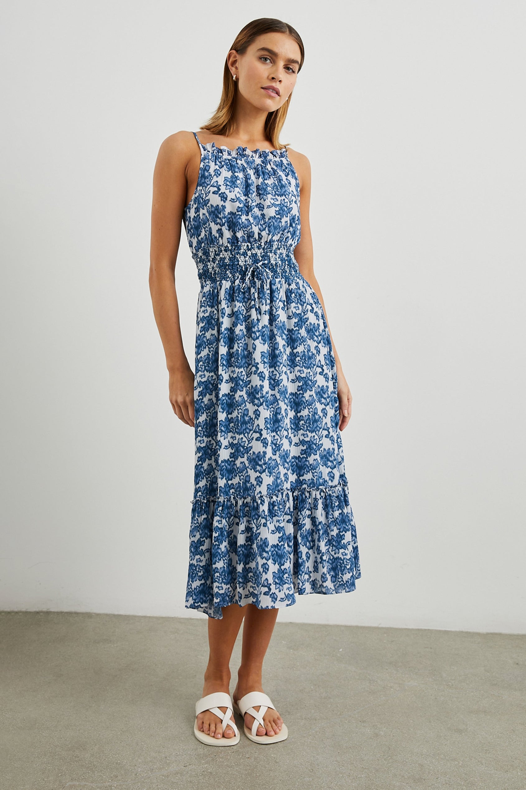 Magdalene Sleeveless Midi Dress with Spaghetti Straps, a Button Keyhole at the Back, a Shirred Waist with a Drawstring, a Scoop Neckline with Ruffled Trim, and a Ruffled Hem in Chambray Floral Colorway, Blue Flowers on White - Front Full Body View Featured on Model  