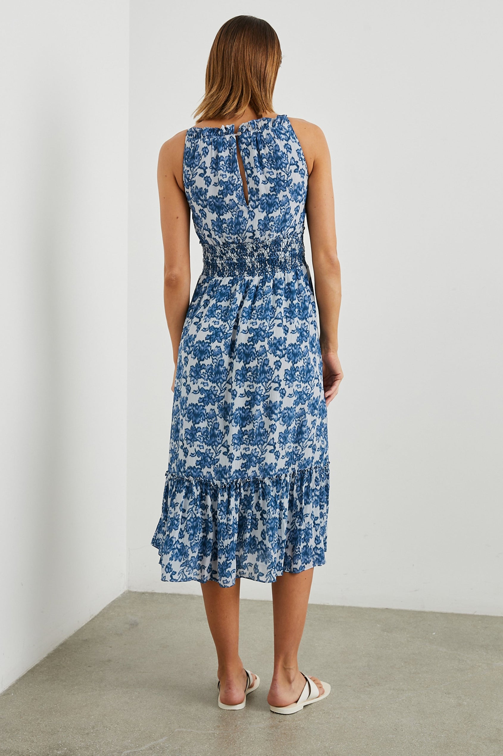 Magdalene Sleeveless Midi Dress with Spaghetti Straps, a Button Keyhole at the Back, a Shirred Waist with a Drawstring, a Scoop Neckline with Ruffled Trim, and a Ruffled Hem in Chambray Floral Colorway, Blue Flowers on White - Back Full Body View Featured on Model  