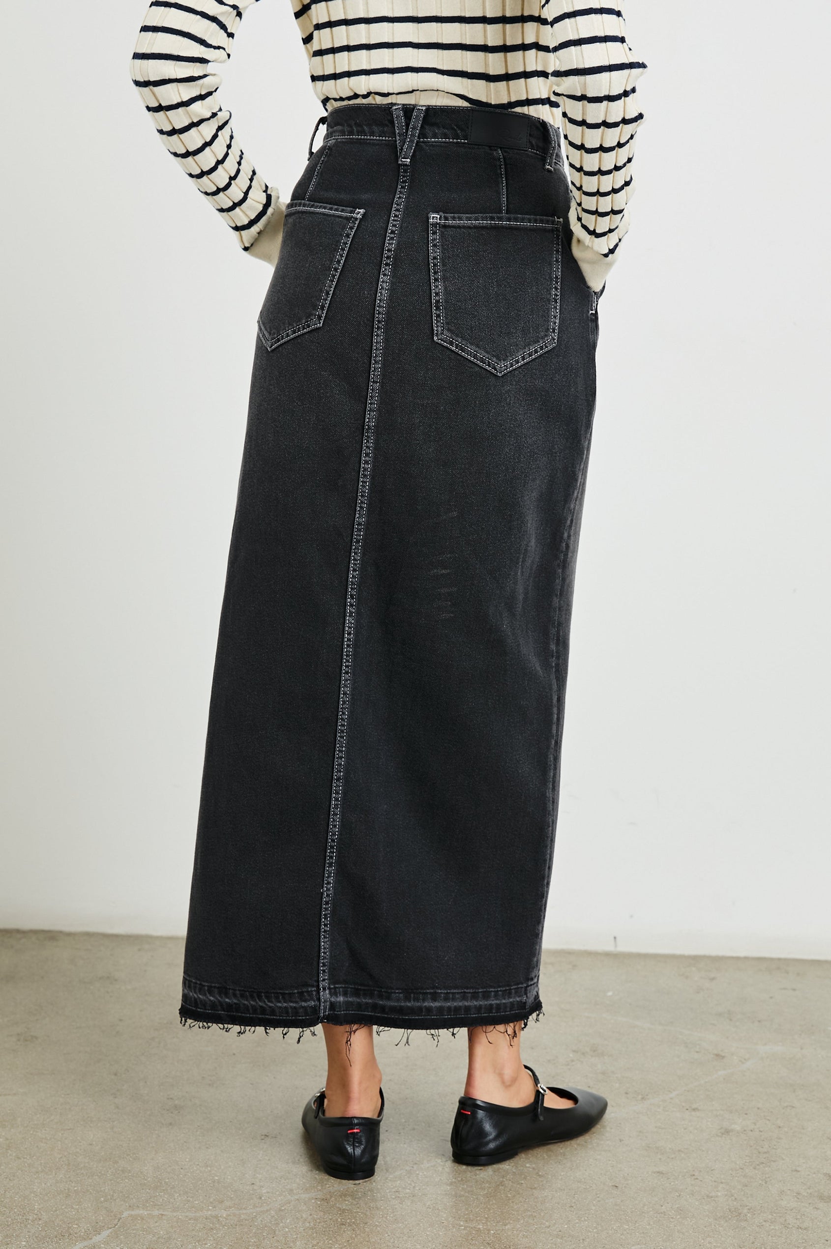 Manhattan Midi Denim Skirt with a Raw Hem, a Front Center Thigh Slit, Belt Loops, Two Front Pockets and Two Back Patch Pockets in Eclipse Colorway, a Black - Back View Featured on Model