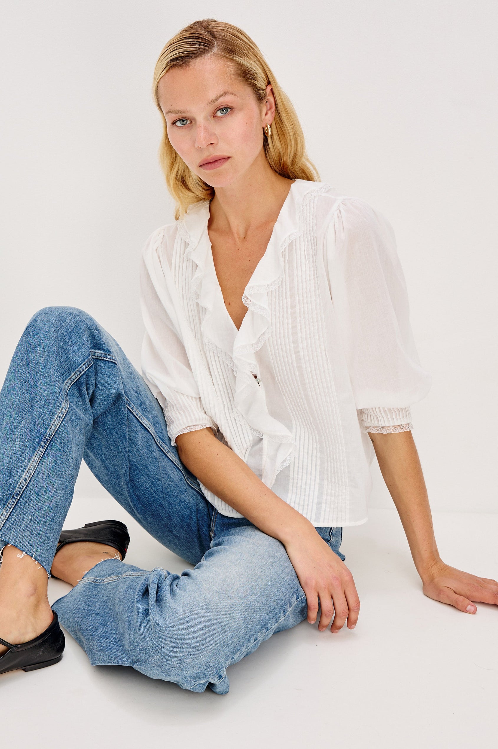 A relaxed-fit cotton blouse with cascading ruffles down the front in White Colorway. The blouse is accented with lace trim at the neckline and cuffs, adding a romantic, feminine touch. Front view with model sitting on the floor.