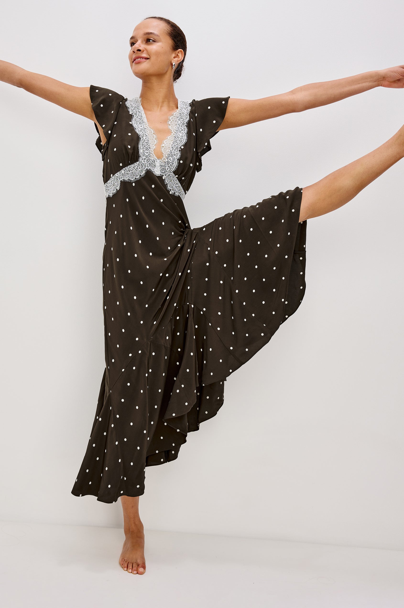 A flowing cocoa polka-dot maxi dress in crepe. The dress has a lace-trimmed V-neckline, ruffle cap sleeves, a hidden side zipper, and godet inserts that add movement to the skirt. Full Body Front View featured on Model gracefully kicking up leg with her arms in the air