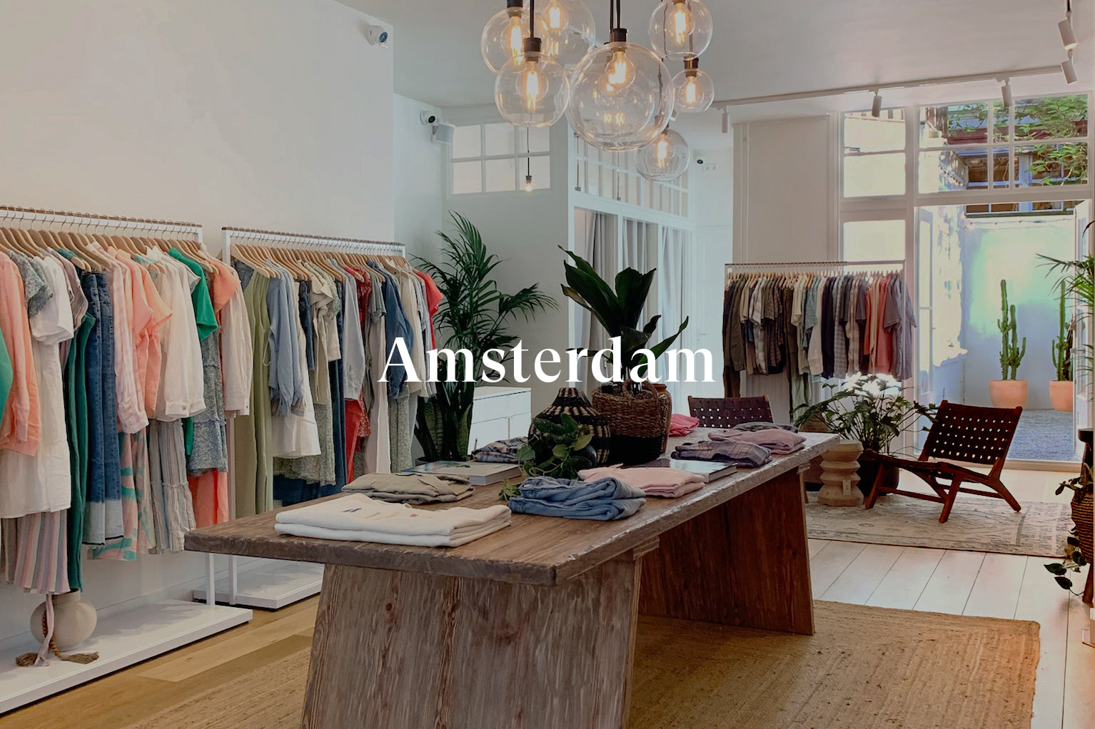 INSIDE OF AMSTERDAM STORE SHOWING LARGE TABLE WITH CLOTHING ON TOP OF IT IN MIDDLE OF ROOM AND CLOTHING RACKS FILLED WITH CLOTHING AGAINST EACH WALL