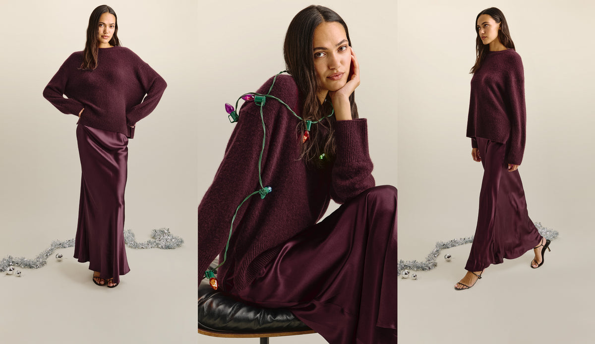 A woman modeling a two-piece outfit consisting of a fuzzy, oversized long-sleeve sweater and a satin-like floor-length skirt, both in a deep plum purple color, set against a plain beige background with silver tinsel on the floor.