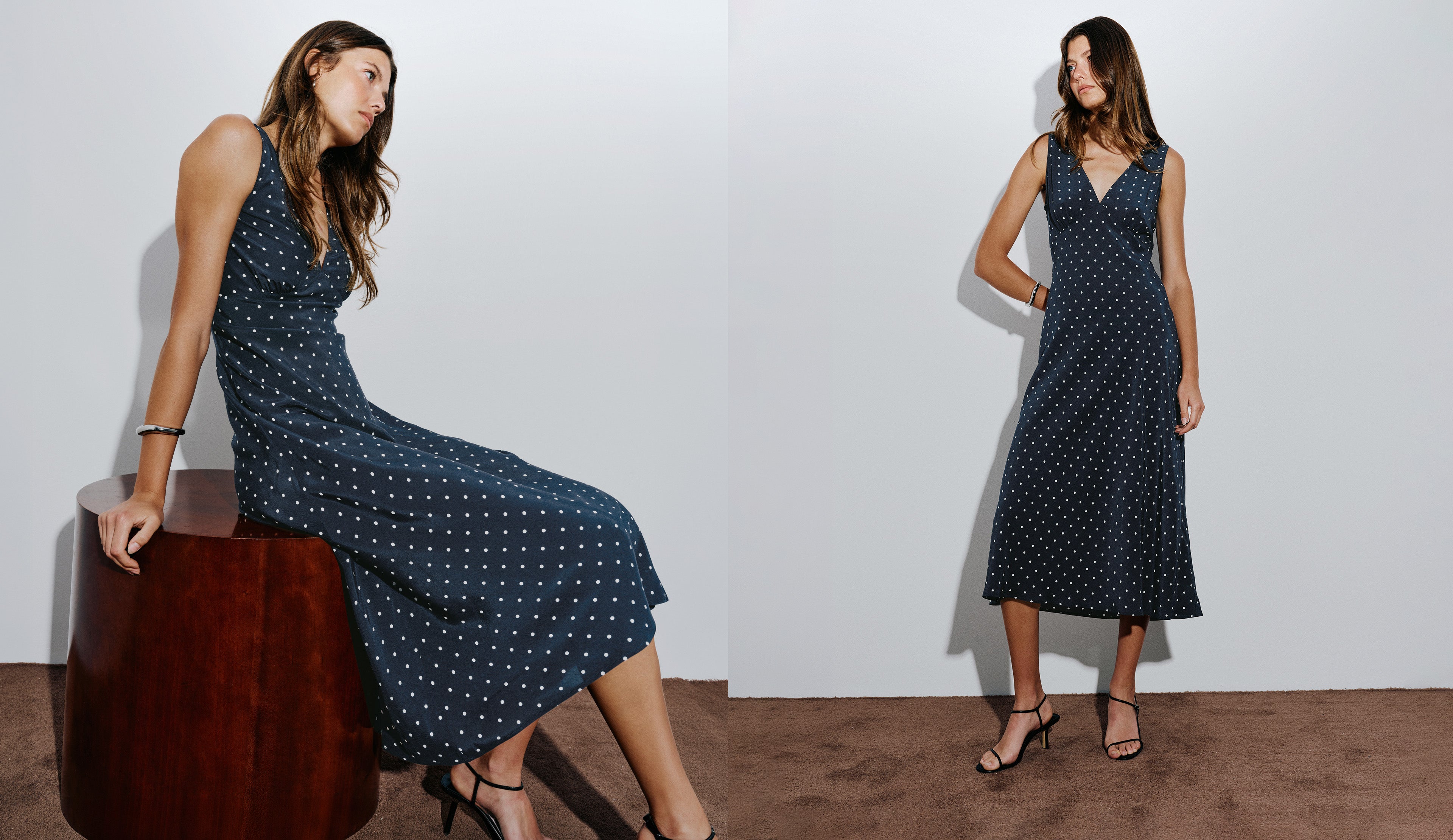 A woman modeling a sleeveless dark navy midi dress with white polka dots, shown in a split composition: seated on a dark wooden stool on the left and standing on the right, against a plain light gray wall.