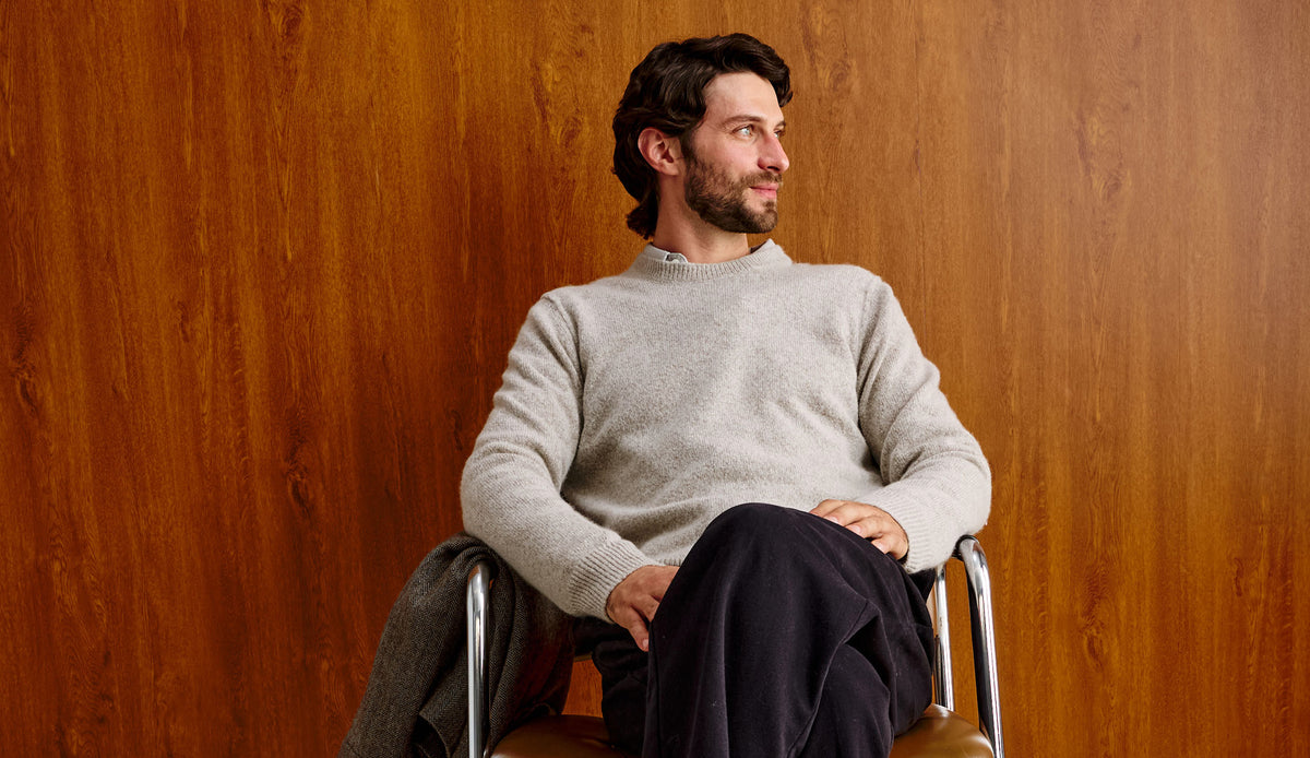 A man with dark hair and a beard sits in a chair against a warm wood-paneled wall, wearing a light gray knit sweater and dark trousers.