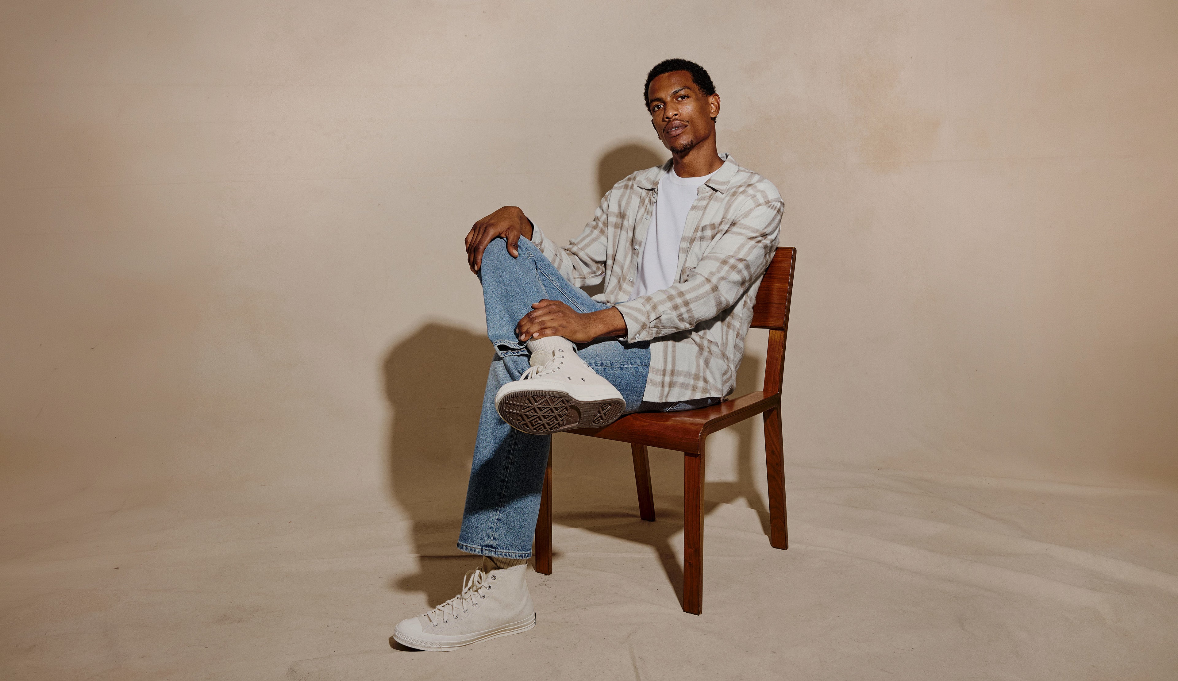 A man sits on a wooden chair against a warm beige studio backdrop, wearing a white and gray plaid button-up shirt over a white t-shirt, blue jeans, and white sneakers.