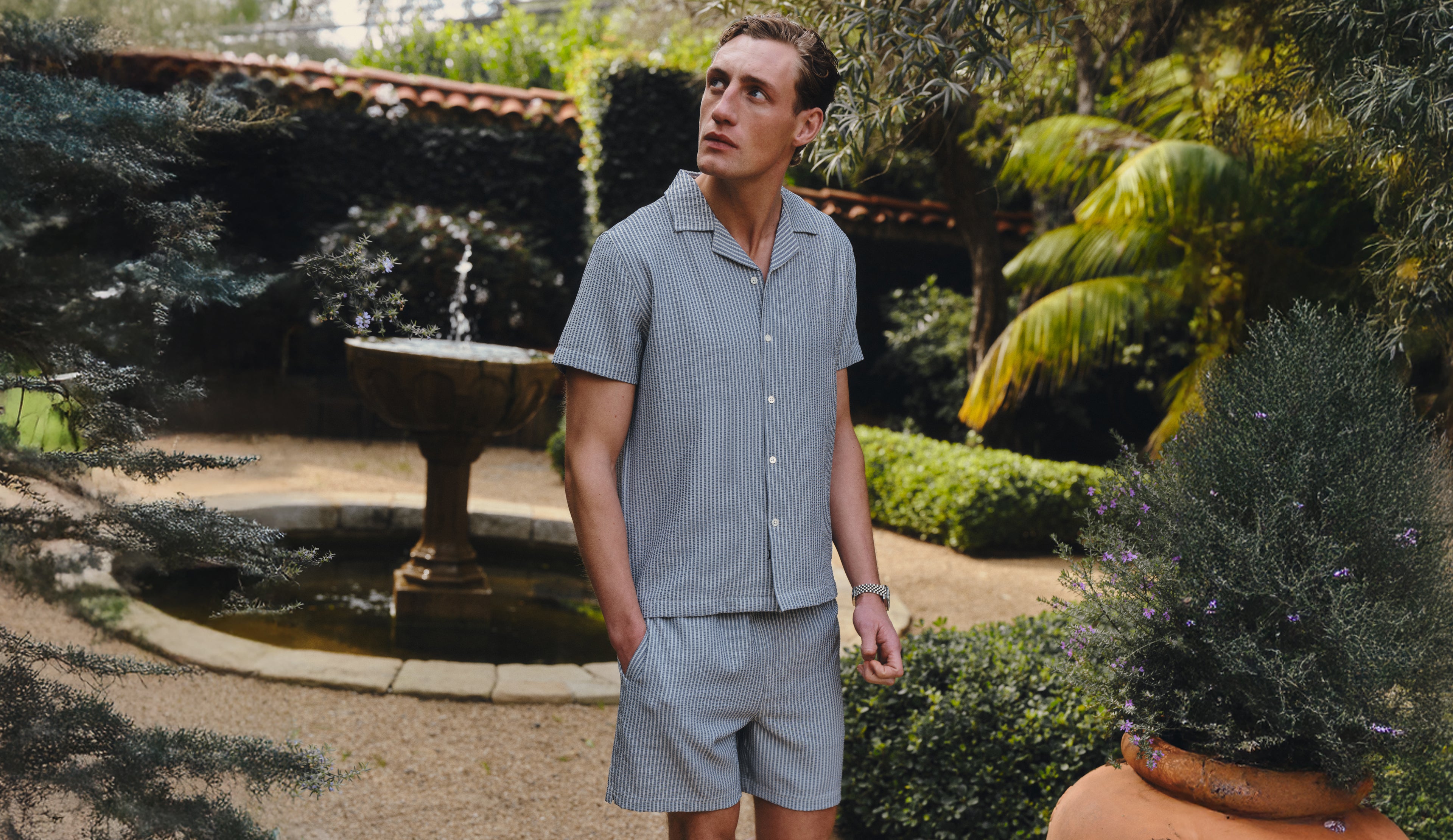 Man in a striped matching set standing in a garden with a fountain and greenery.