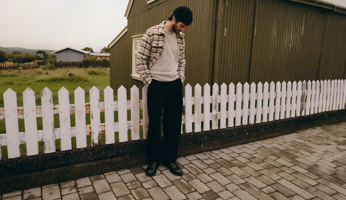A man wearing a black beanie, a brown and white plaid flannel shirt over a white crewneck sweater, black trousers, and black shoes stands on a paved path next to a white picket fence and a dark green barn.
