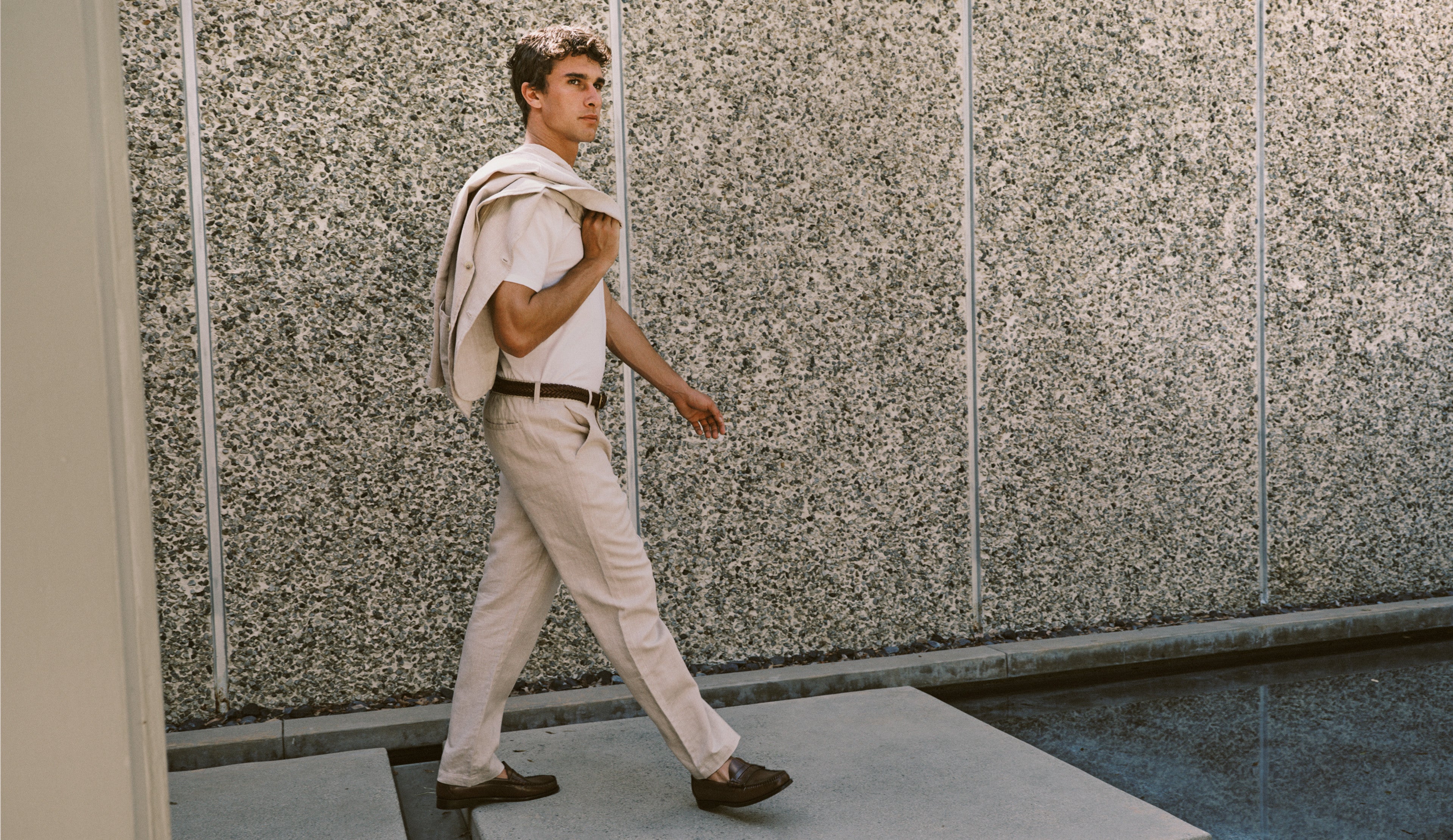A man walking outdoors wearing a light beige short-sleeve shirt, matching beige trousers, and dark brown loafers, with a light-colored jacket draped over his shoulder, set against a textured concrete wall.