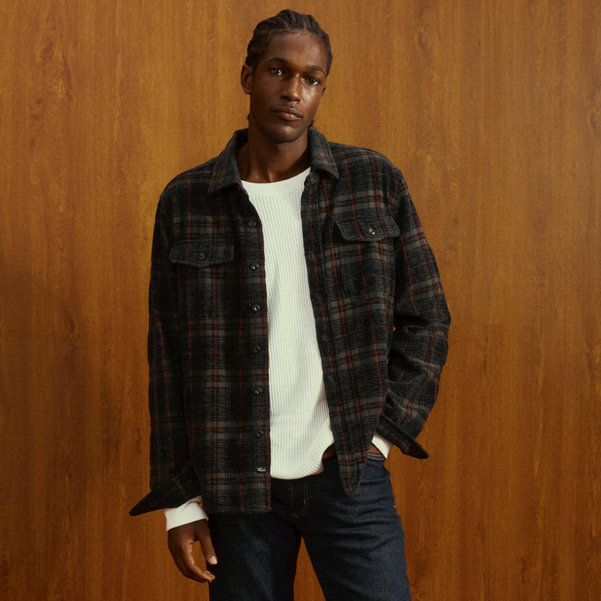 A man wearing a dark plaid button-up overshirt over a white ribbed sweater and dark jeans, standing against a warm wood-paneled background.