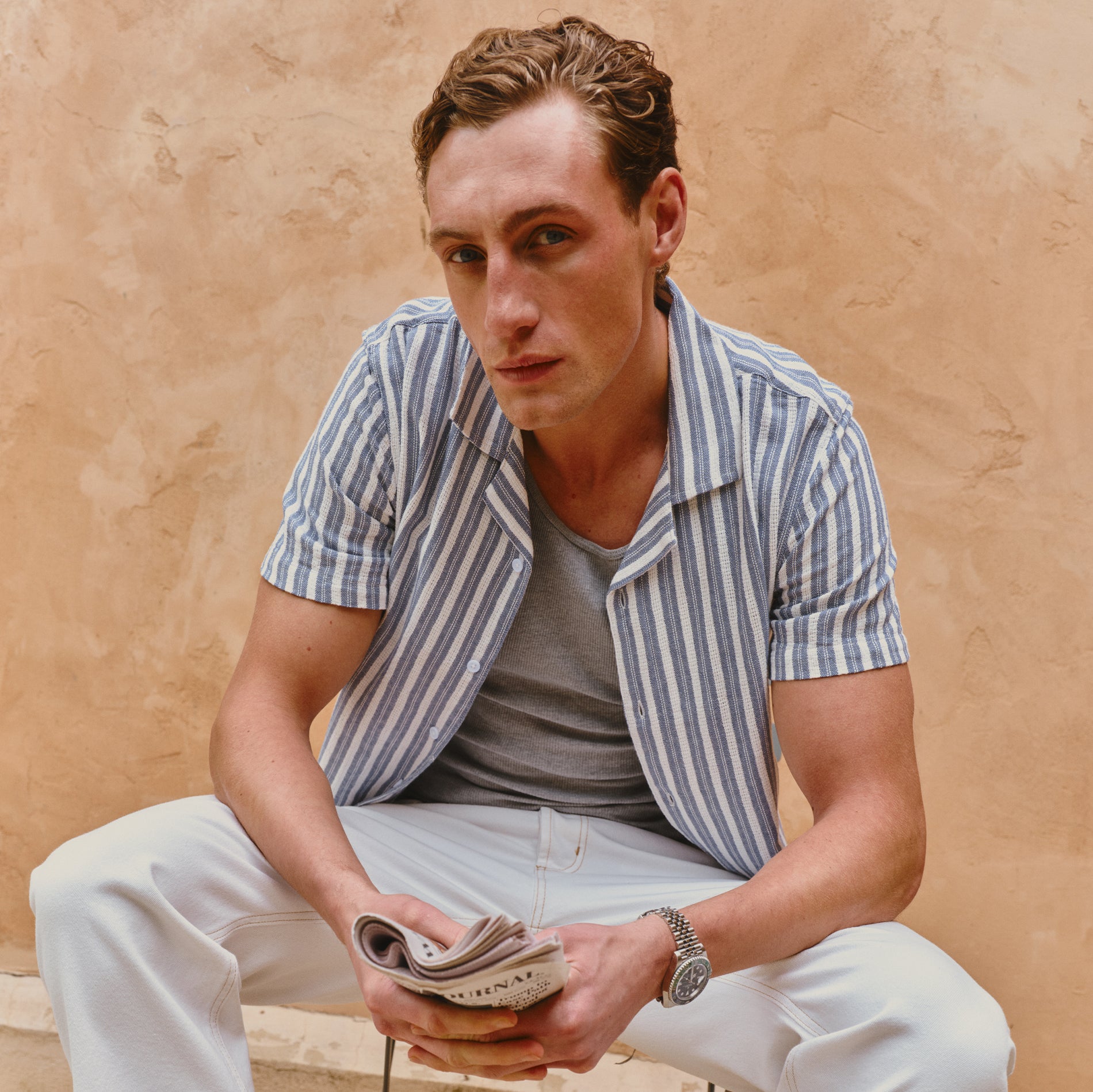 Man sitting against a beige wall wearing a striped shirt and holding a newspaper.