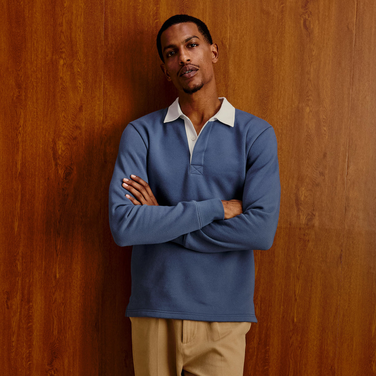 A man wearing a slate blue long-sleeve rugby-style polo shirt with a white collar, standing with arms crossed against a warm wood-paneled background.