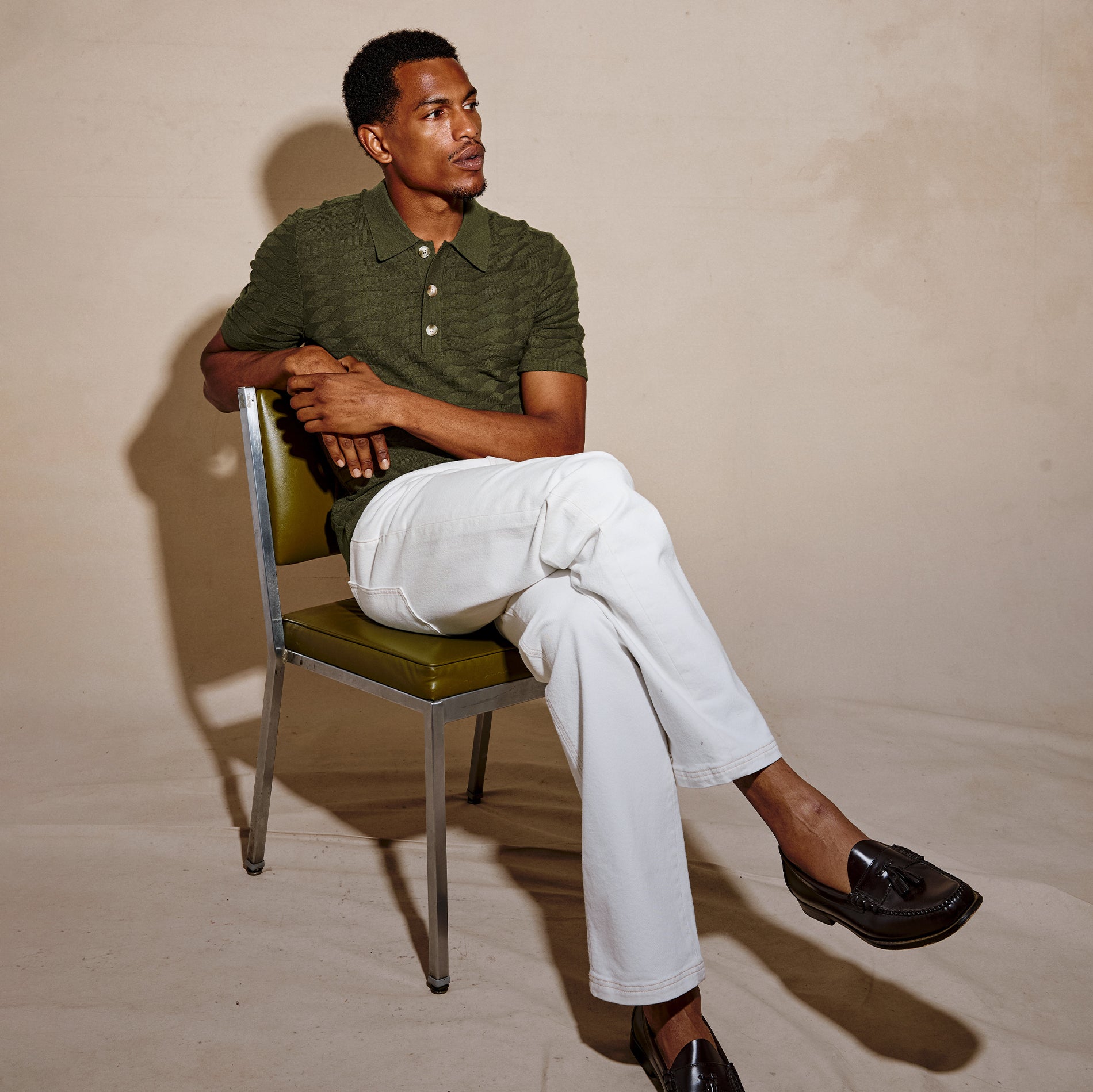 A man sits on a green and chrome chair wearing a dark olive green striped polo shirt, white trousers, and black loafers against a beige studio backdrop.