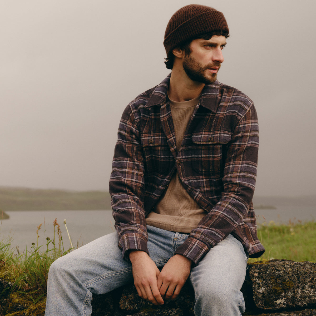 A man wearing a brown knit beanie, a brown and black plaid flannel shirt over a beige t-shirt, and light blue jeans sits on a stone wall outdoors with a misty lake and grassy hills in the background.