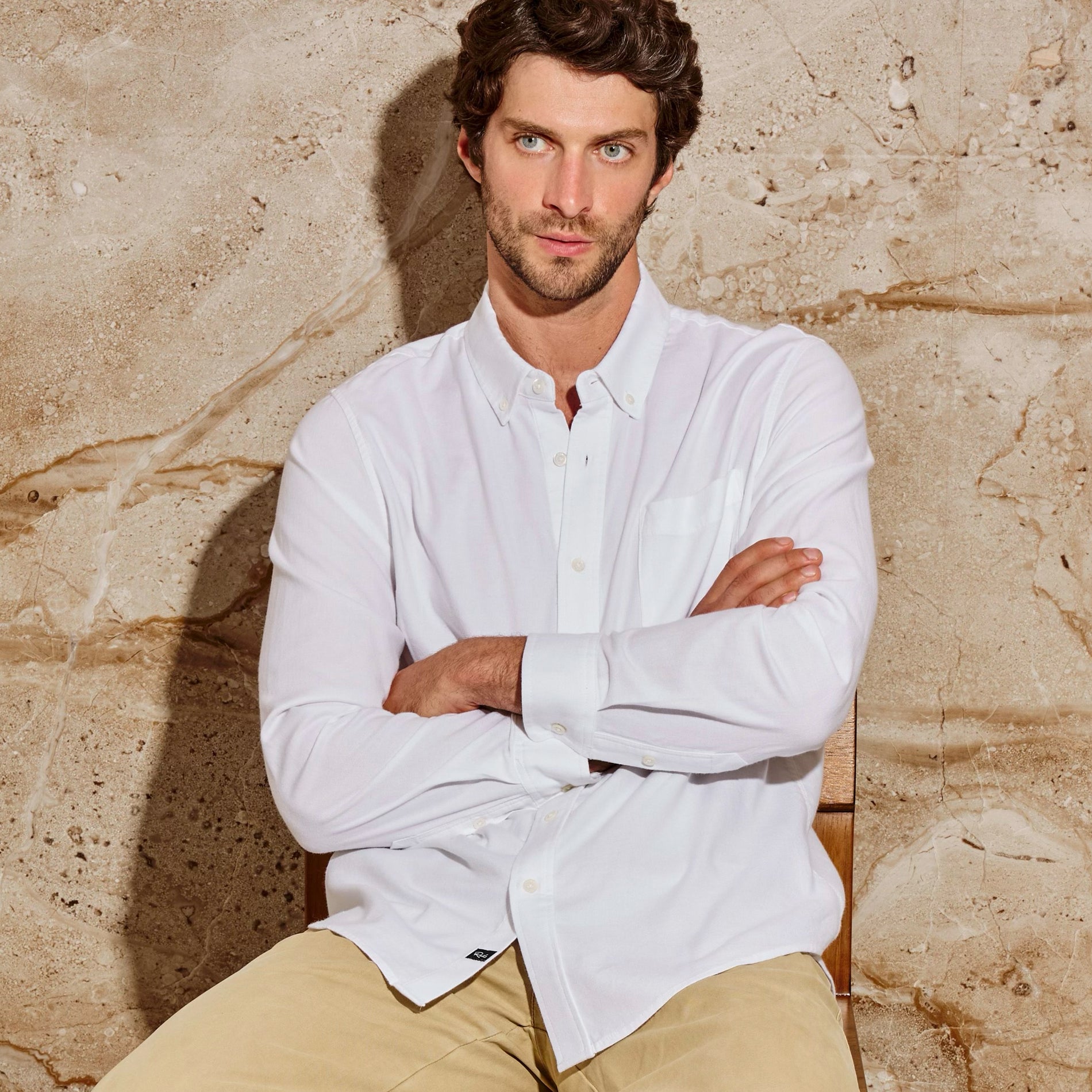 Man wearing a white shirt and beige pants sitting against a stone wall.